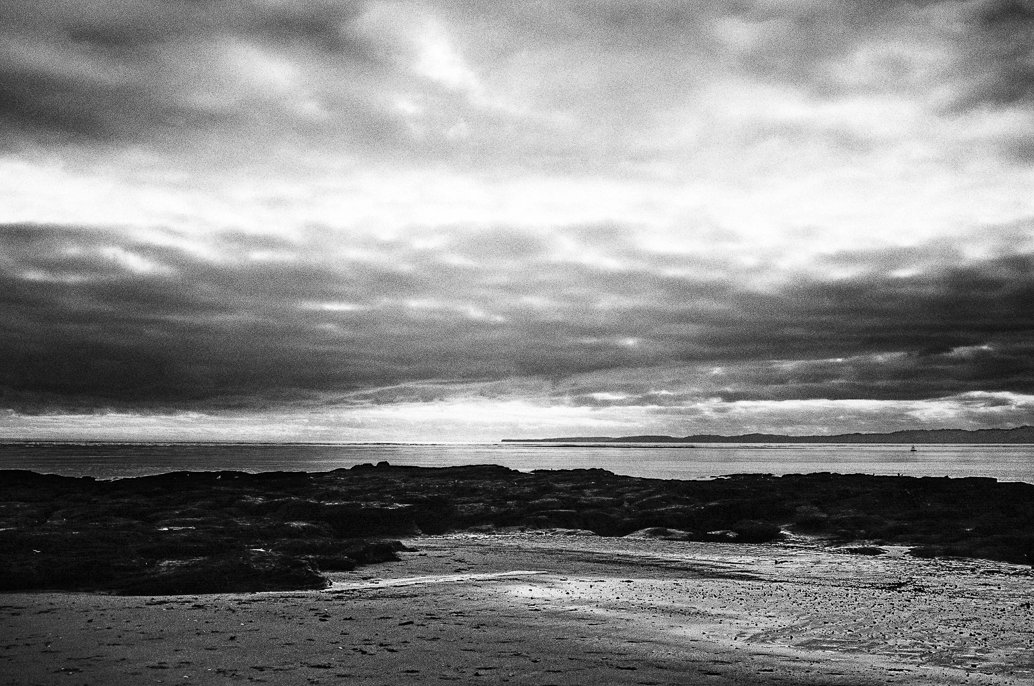Black and white photo of a cloudy sky over a rocky beach with the ocean in the background and distant land across the water.