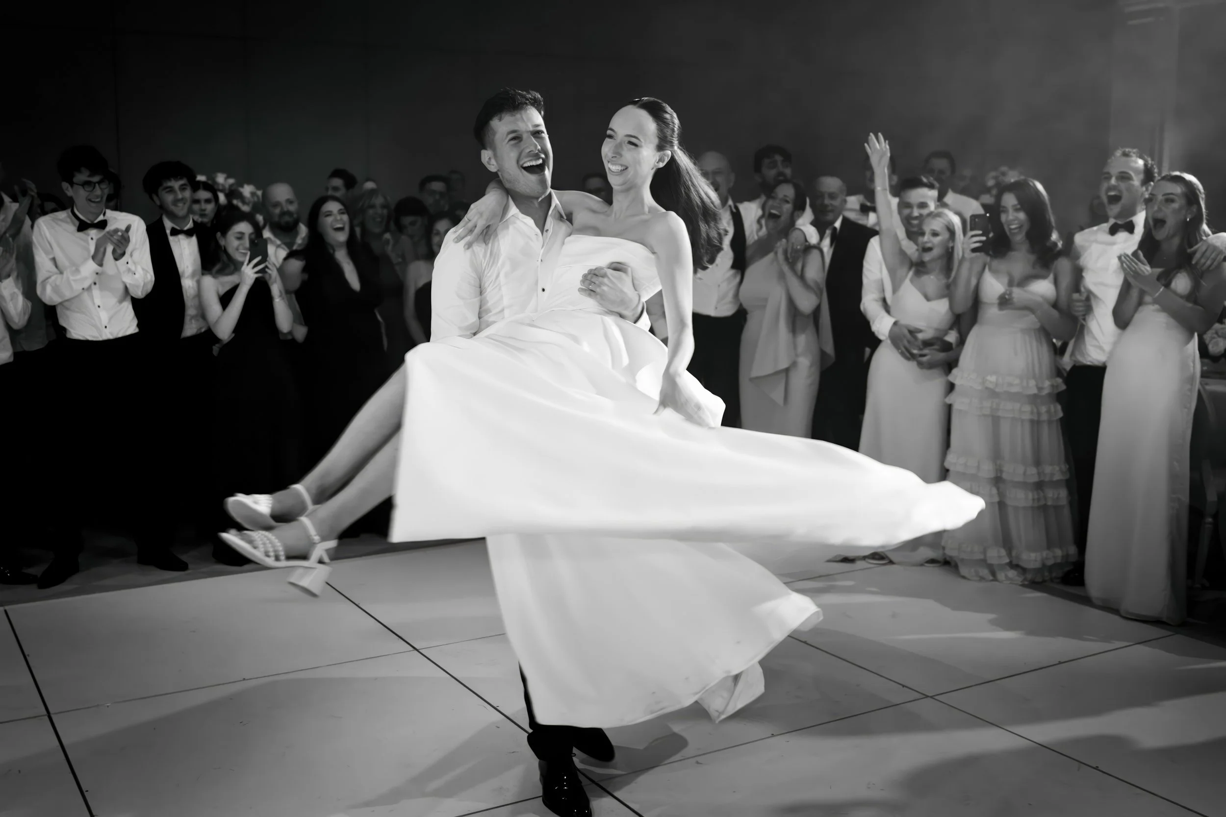 A groom lifting the bride during their wedding reception, surrounded by guests clapping and cheering in a large indoor venue.