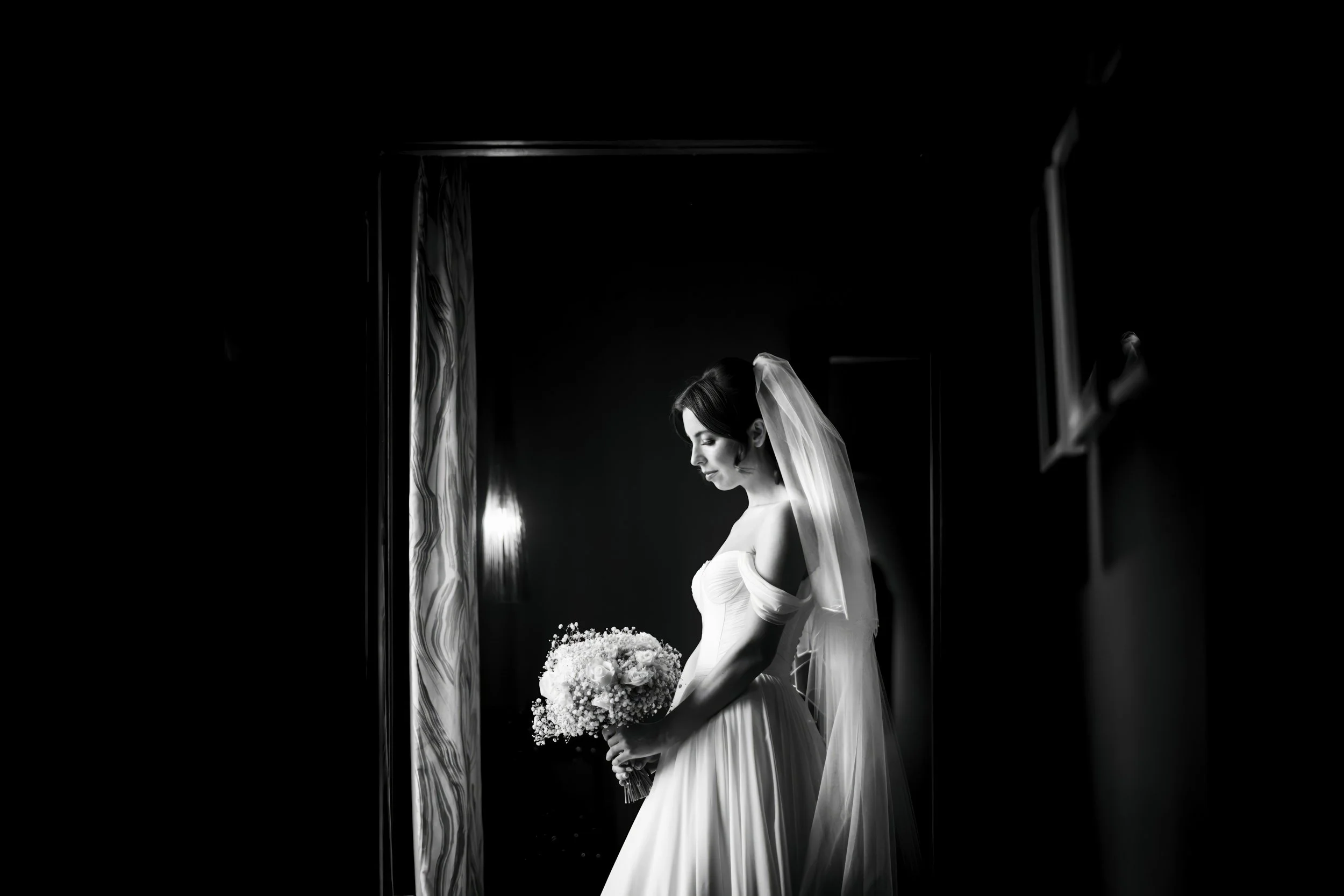 Black and white photo of a bride in a wedding dress holding a bouquet, standing in profile in a dimly lit room with curtains and a window.