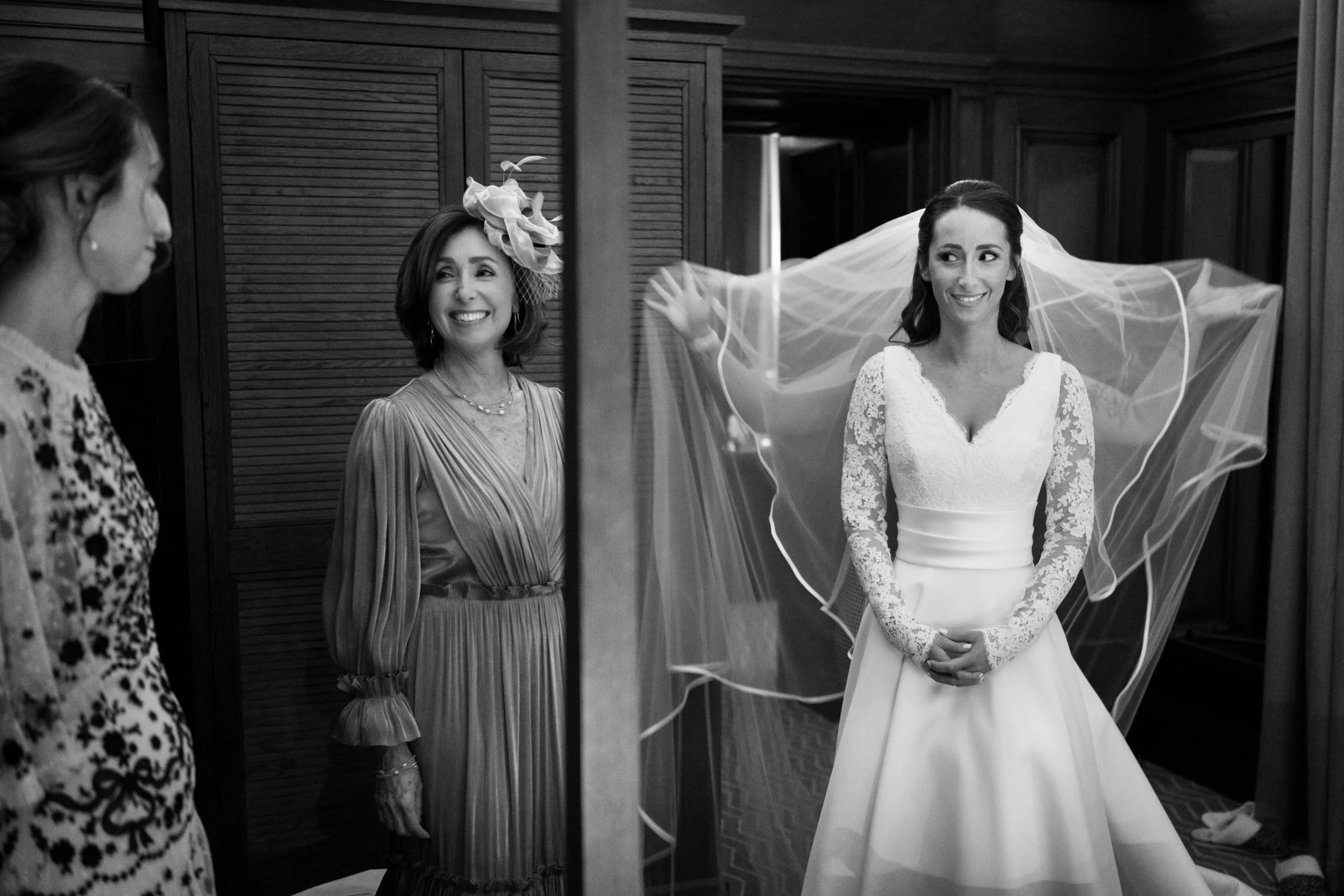 A bride in a wedding dress with lace sleeves smiling at her mother and another woman in a patterned dress, who are looking at her.