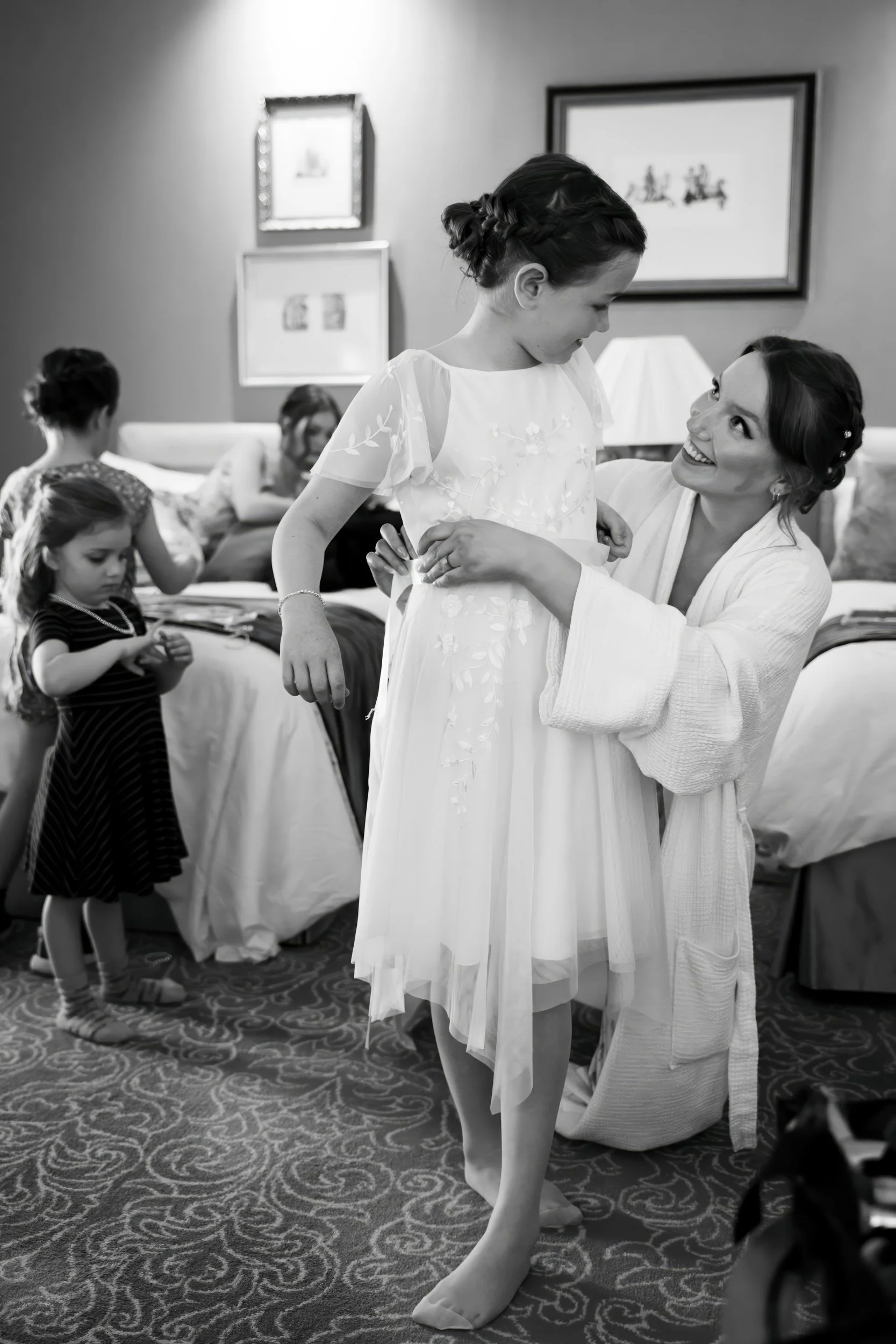 A woman helping a young girl in a white dress, smiling and looking at each other in a hotel room. In the background, three other young girls are engaged in different activities.
