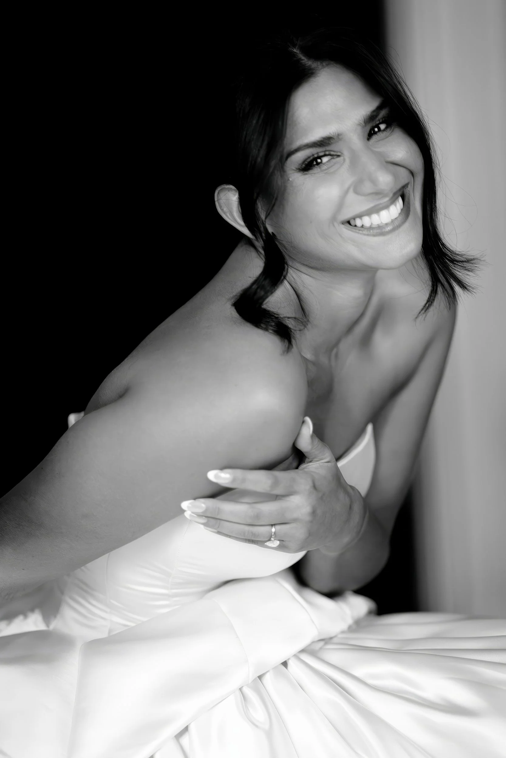 Black and white photo of a smiling woman with dark hair, wearing a strapless dress, holding her left arm across her chest, and sitting on her lower body with a satin fabric.