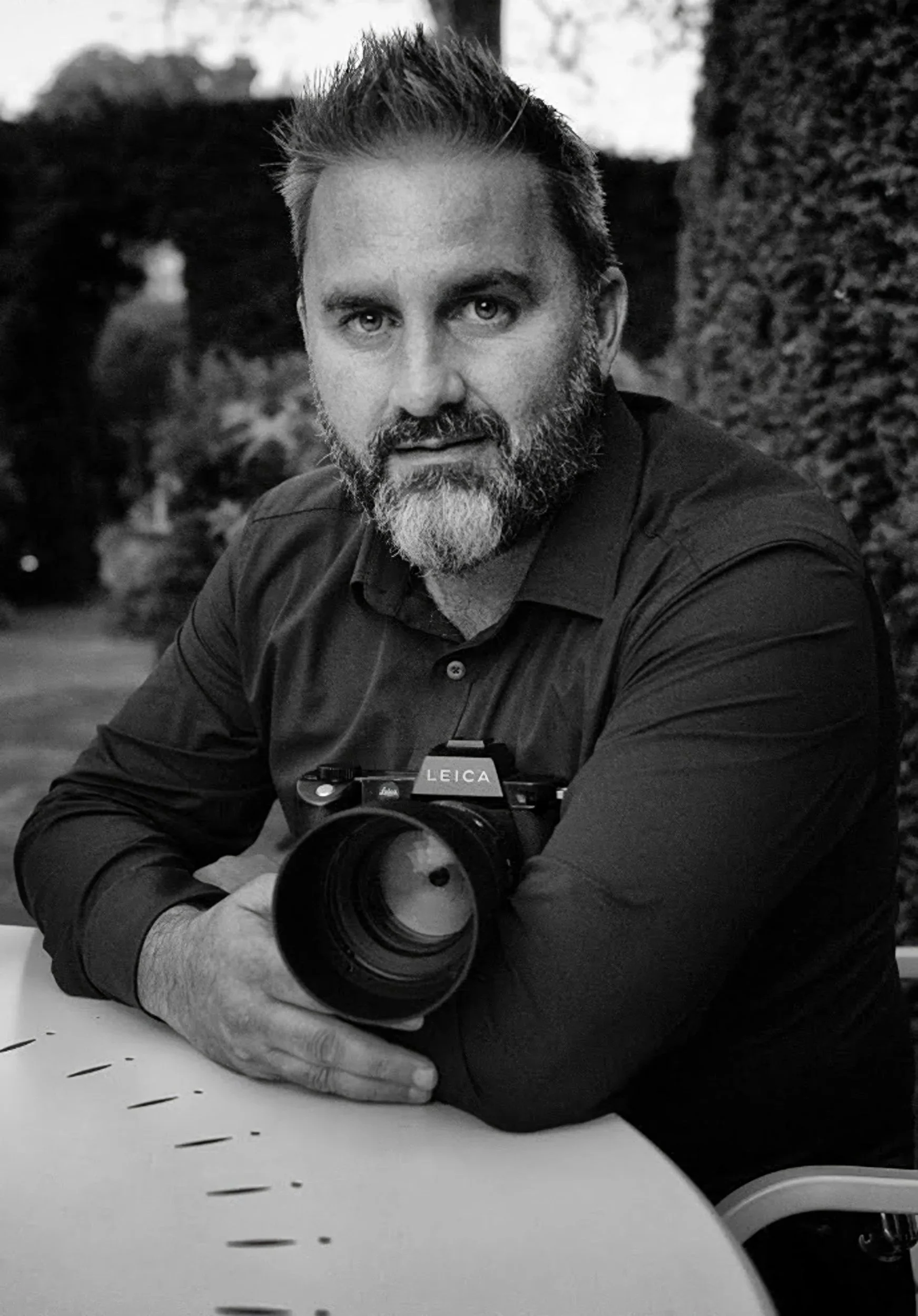 A man with a beard sitting outdoors, holding a Leica camera, looking at the camera, with trees and bushes in the background.