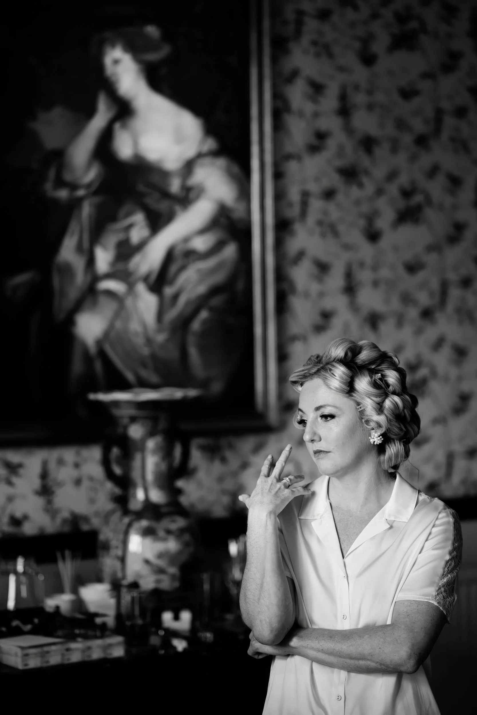 A woman with styled hair and earrings, wearing a satin blouse with lace sleeves, stands in front of a large, classical portrait painting in an art gallery or museum, with her hand touching her face thoughtfully.