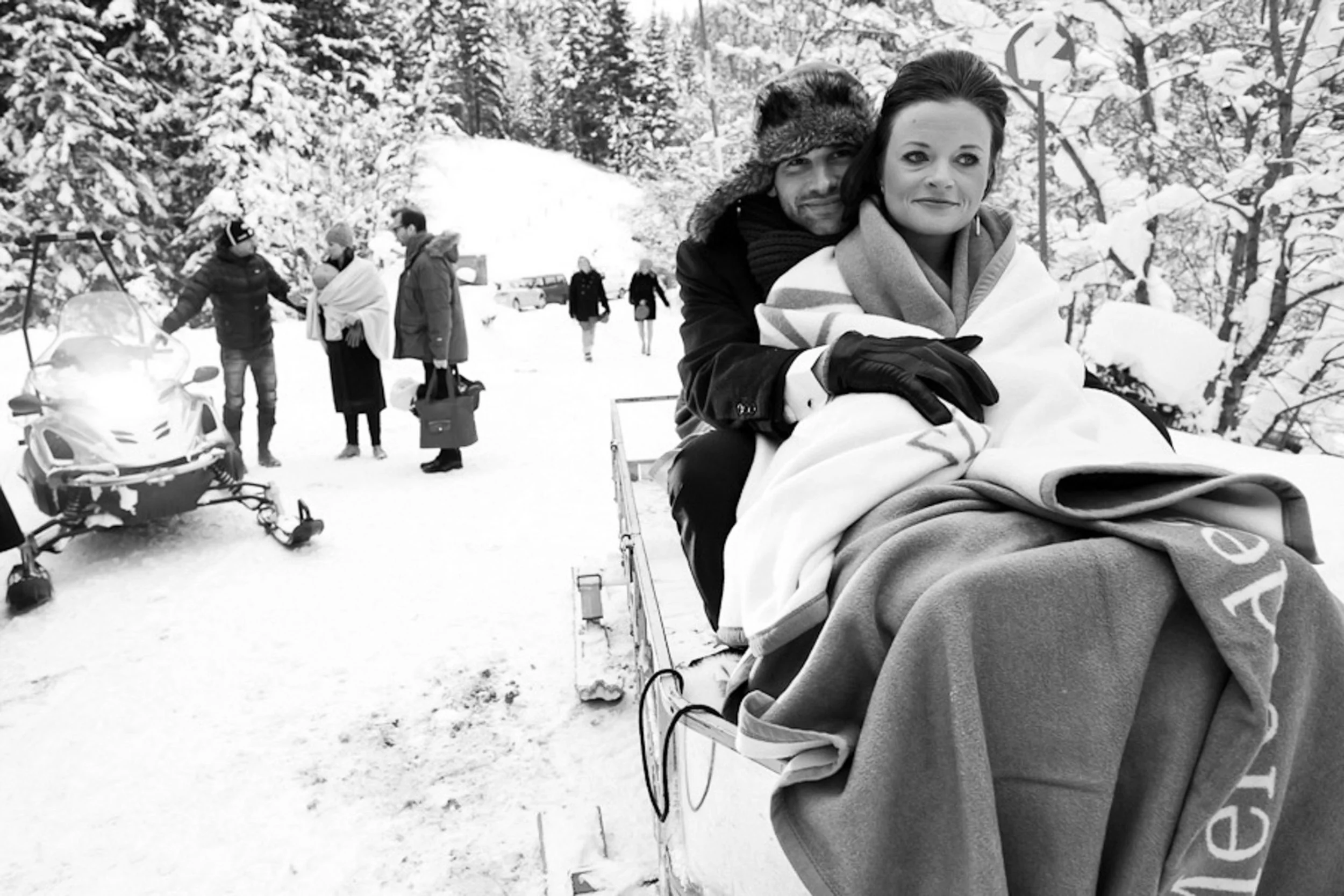 A man and woman sitting on a sled in snowy woods; the man is wearing a fur hat and gloves, and the woman is wrapped in blankets. In the background, people are standing near snowmobiles and walking through the snow, with snow-covered trees around.