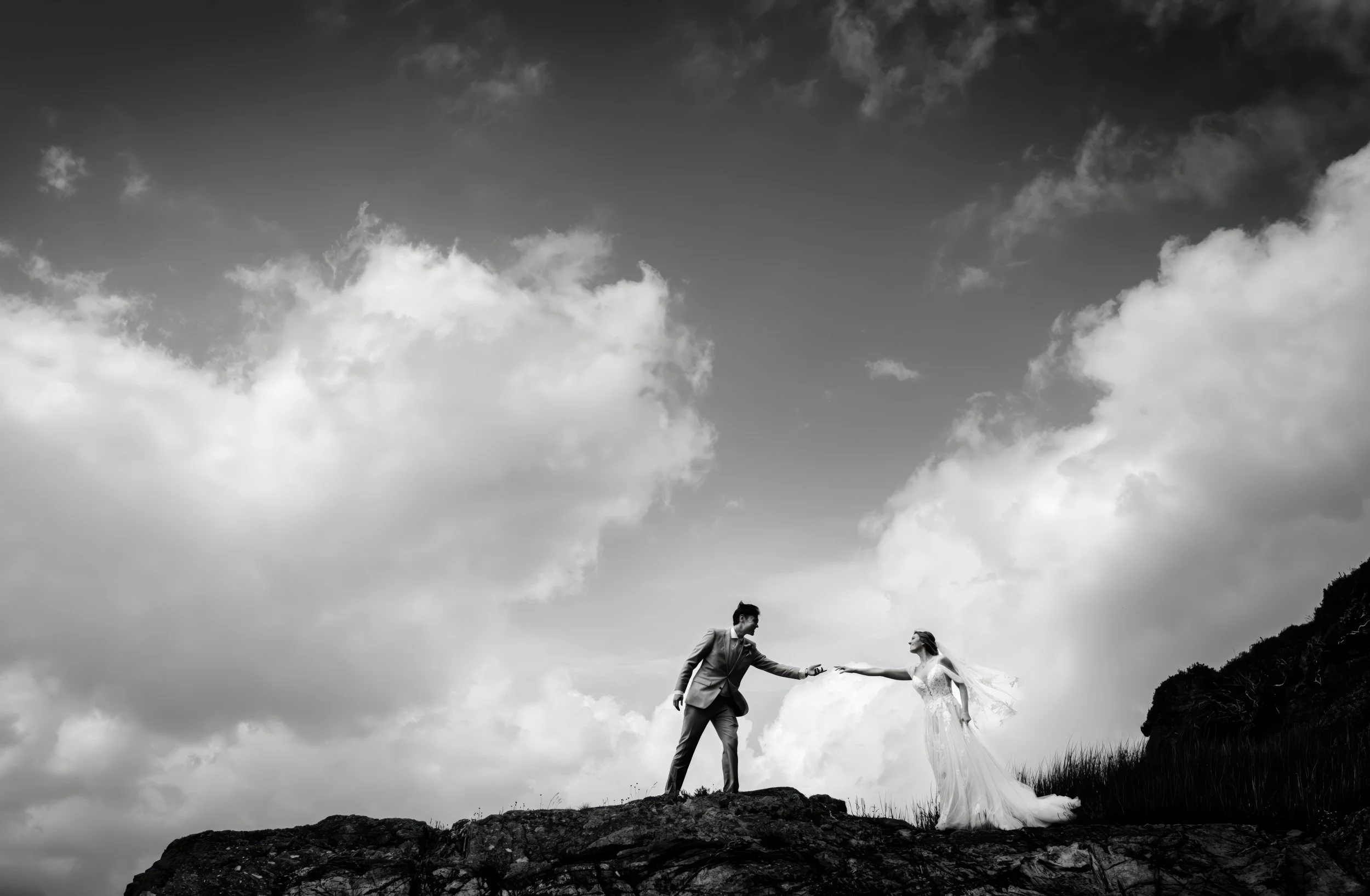 Black and white photo of a man in a suit reaching out his hand to a woman in a wedding dress on a rocky hill with a cloudy sky background.