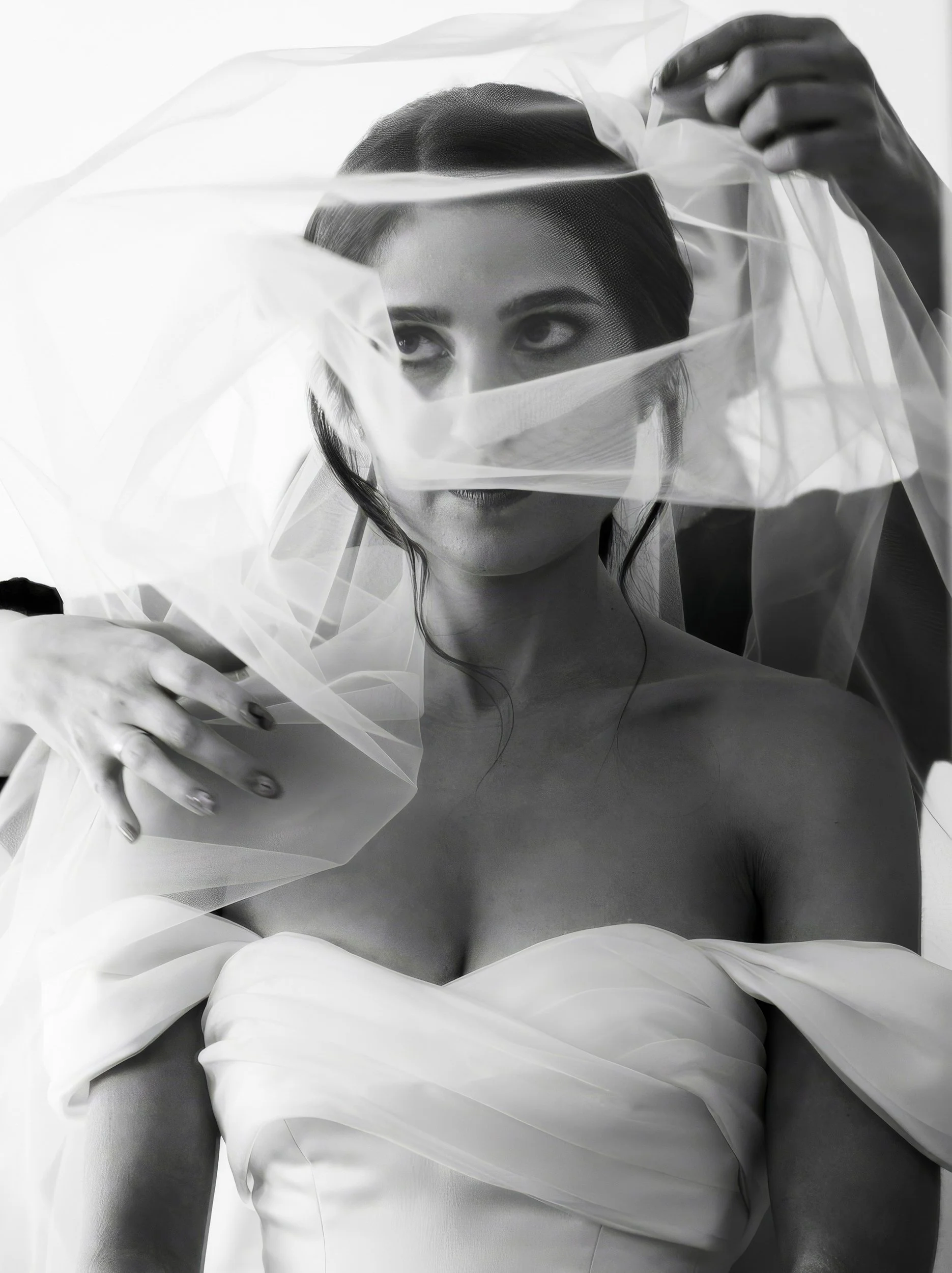 A black and white photo of a woman with short hair, wearing a strapless dress and holding a sheer fabric veil over her face, with her eyes visible through the fabric.