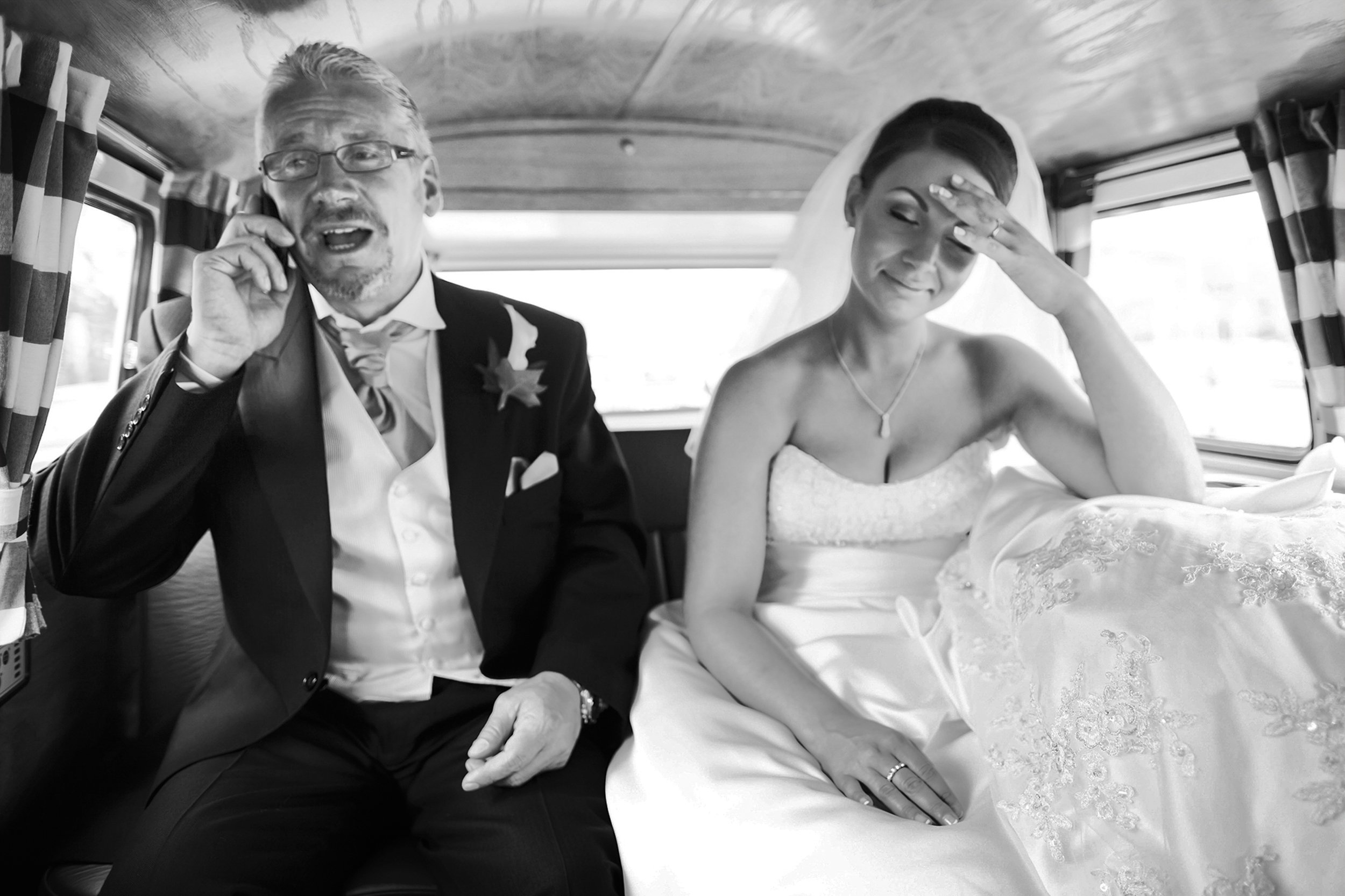 A groom in a tuxedo talking on a phone and a bride in a wedding dress sitting next to him, inside a vehicle with patterned curtains.