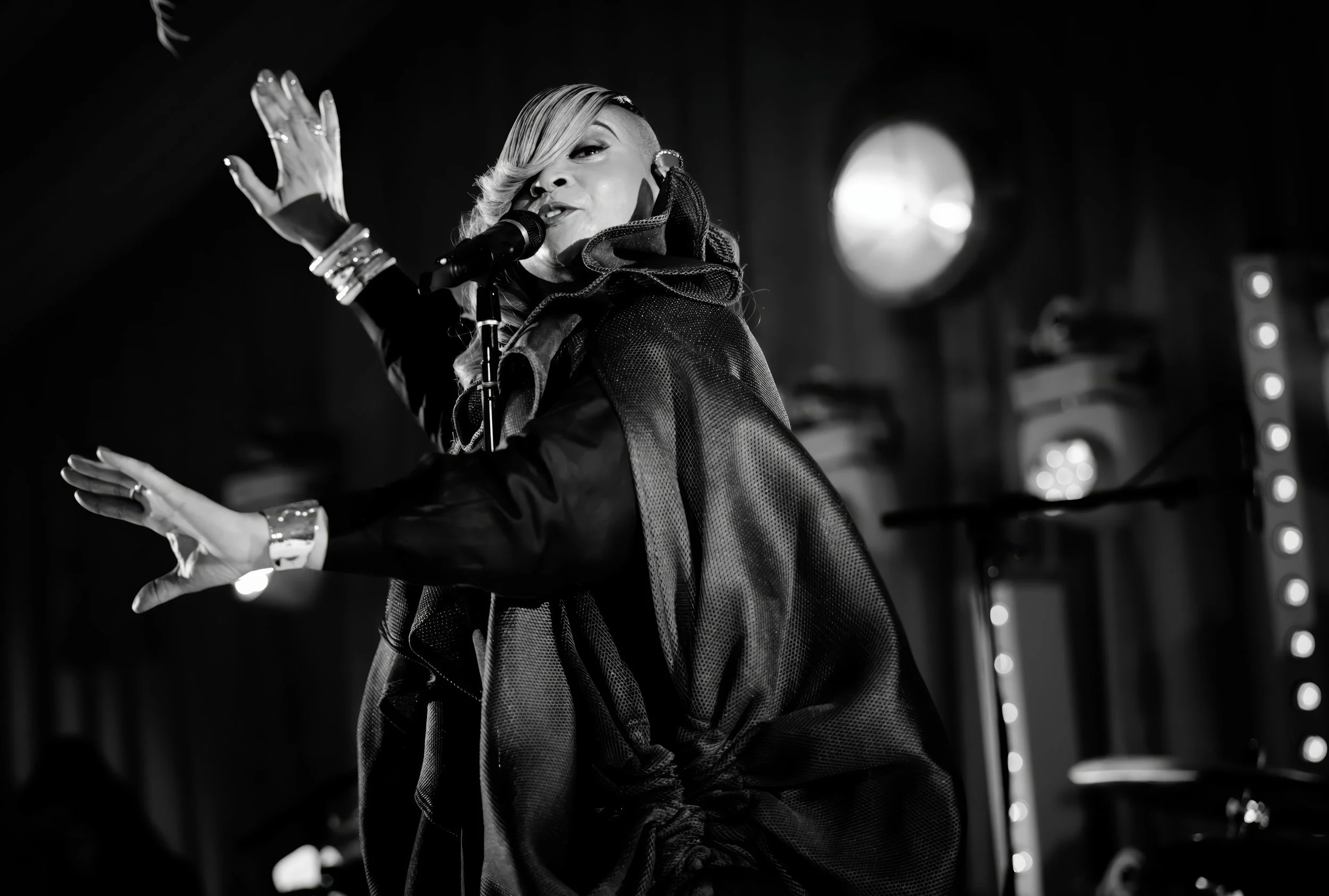 A female singer performing on stage with a microphone, black and white photo, wearing a dark jacket with a large collar, her hair styled in a side swept look, stage lights behind her.