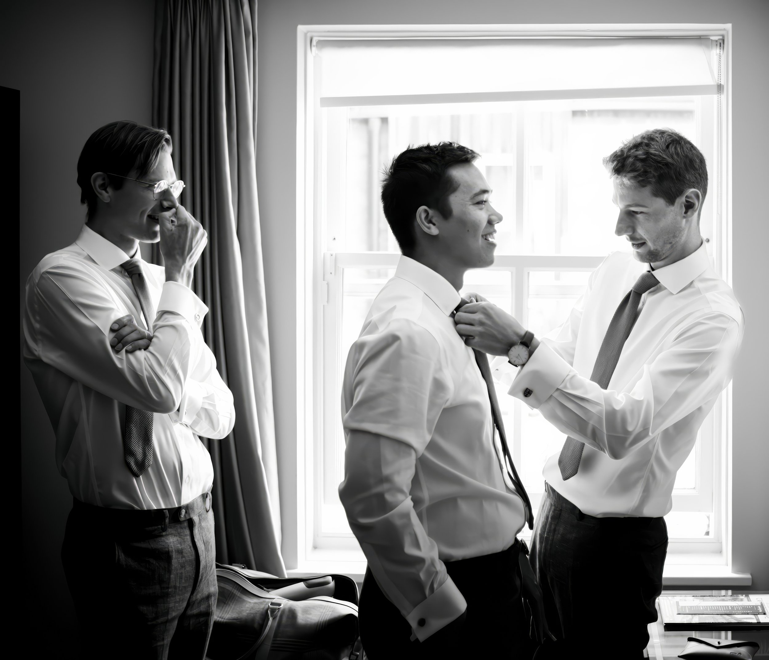 Three young men in business attire in a room with a large window. One stands in the center, smiling, while another adjusts his tie. The third stands to the side, laughing, with hand over mouth.