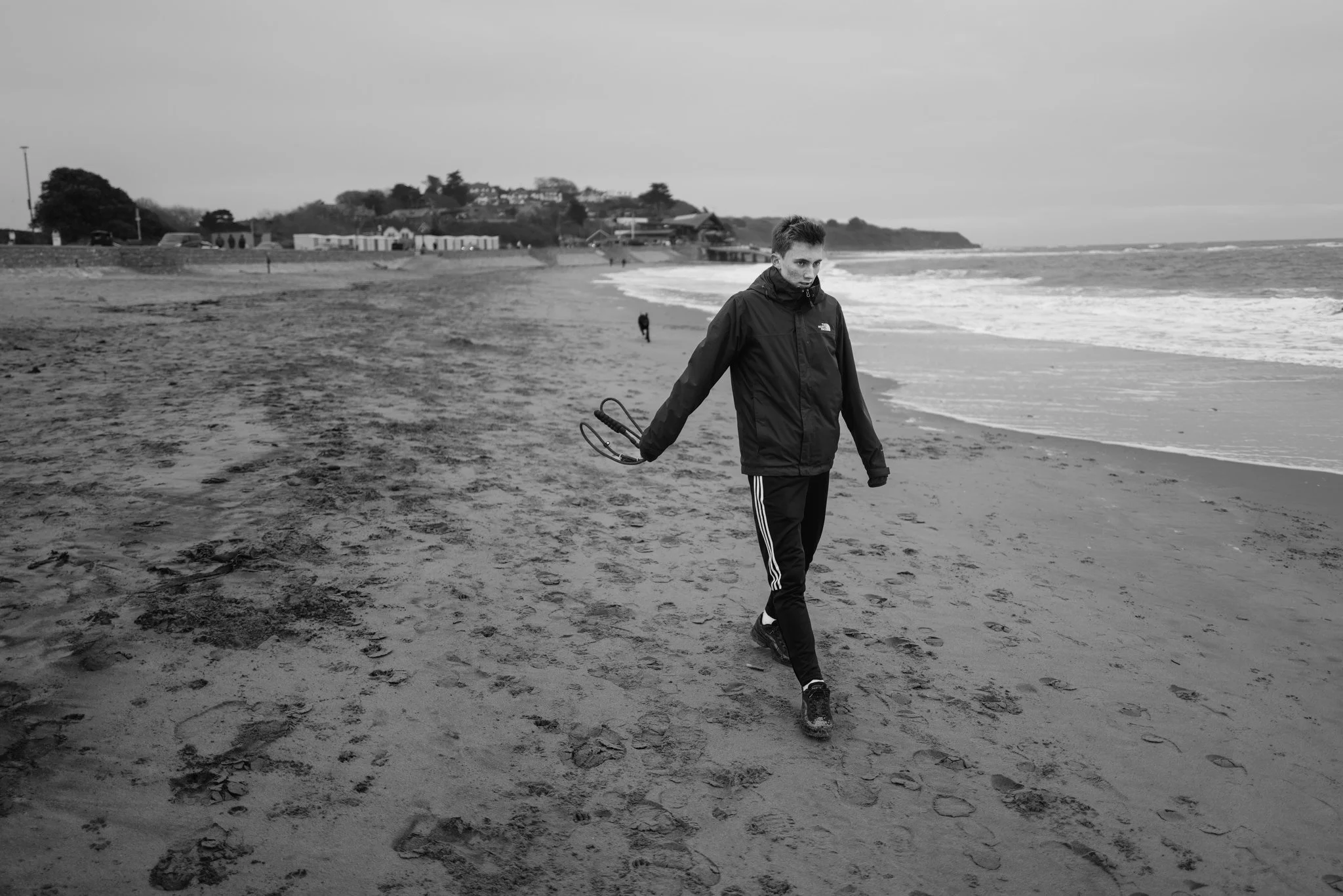 A young man walks along a deserted beach on a cloudy day, holding sunglasses in his right hand, wearing a dark jacket and track pants.