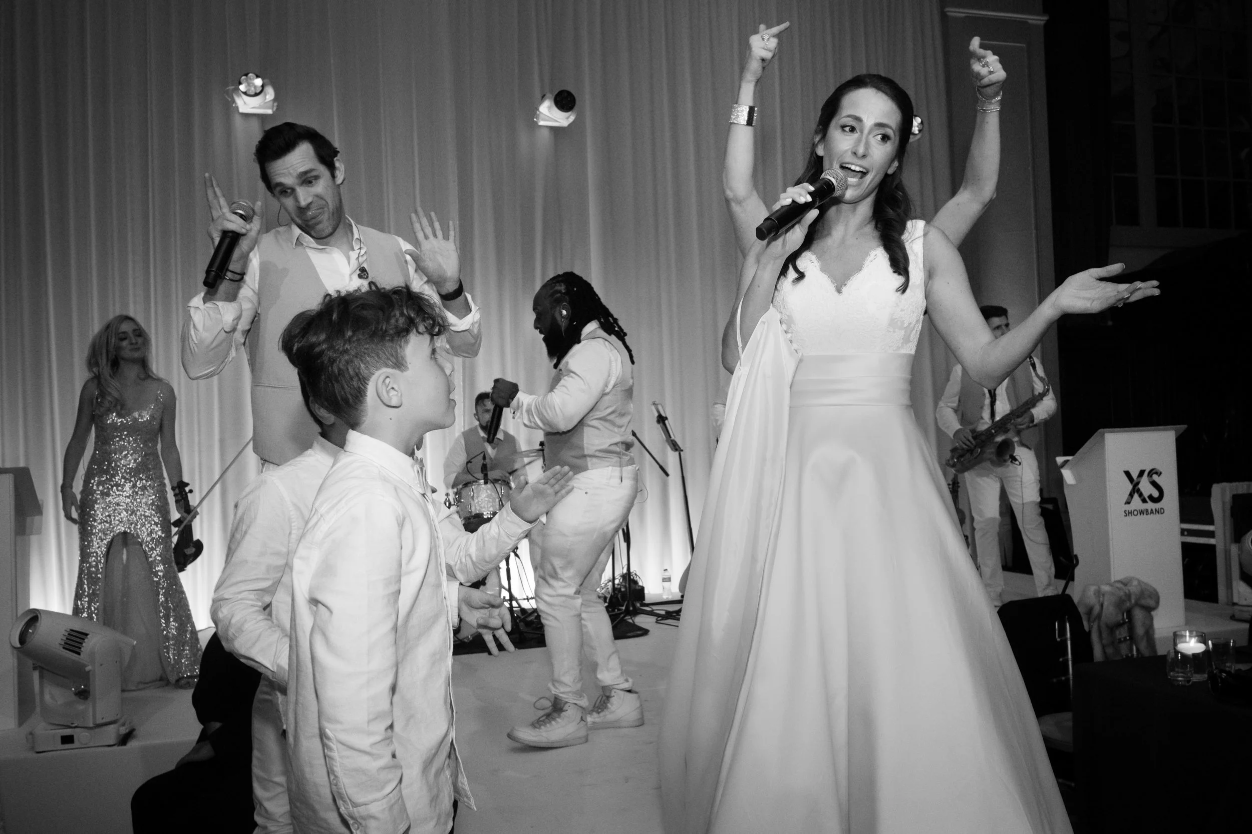 A wedding reception with the bride singing into a microphone, guests dancing, and a band performing on stage in the background.