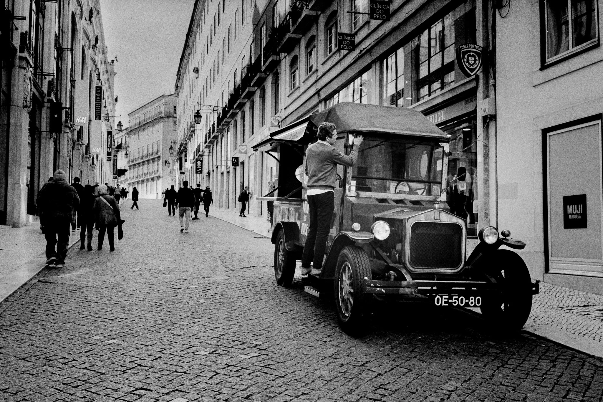 A vintage car with a man leaning on it on a cobblestone city street, with pedestrians walking in the background.