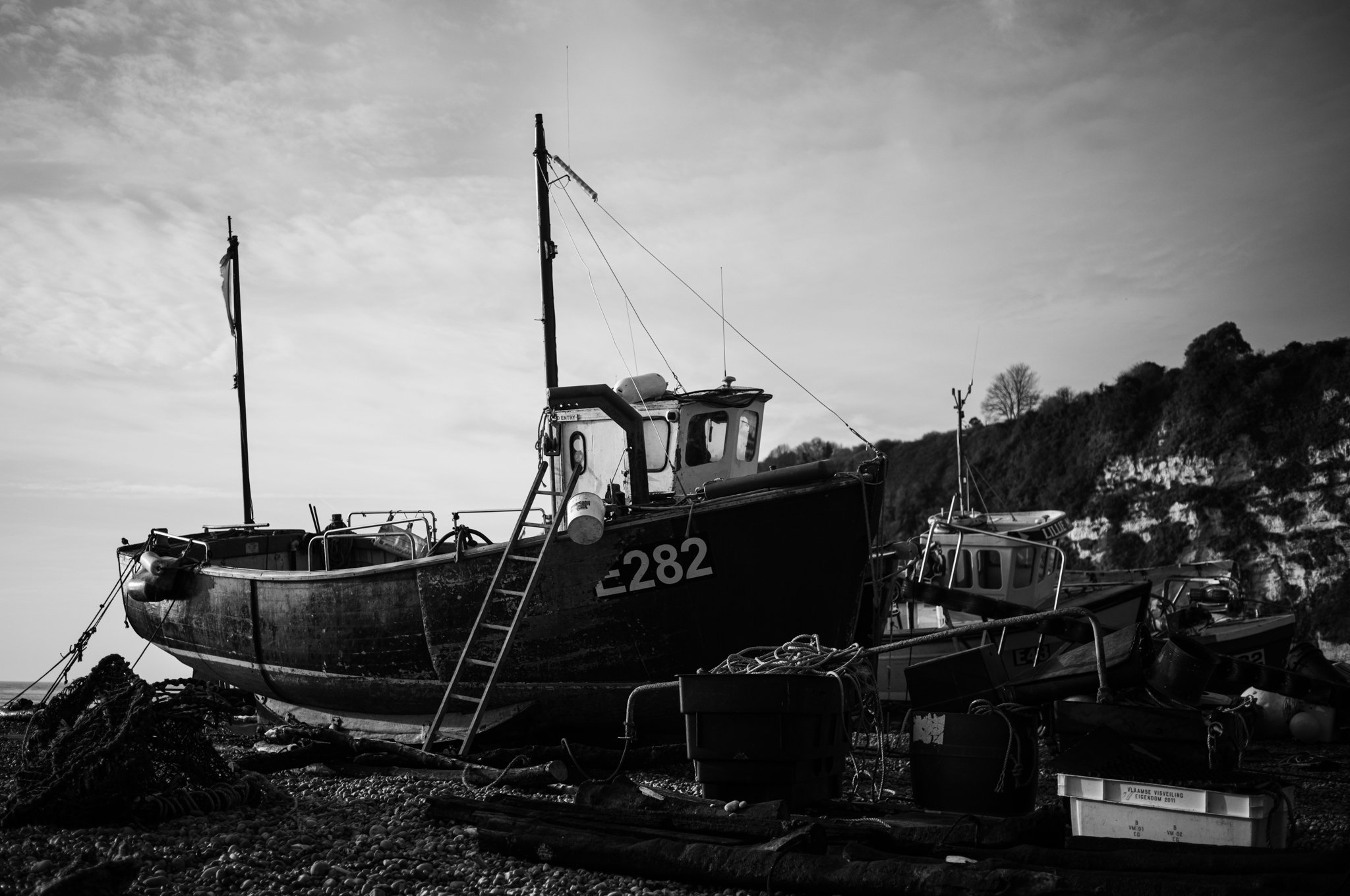 Black and white photo of anchored boats on rocky shore with a hill in the background.