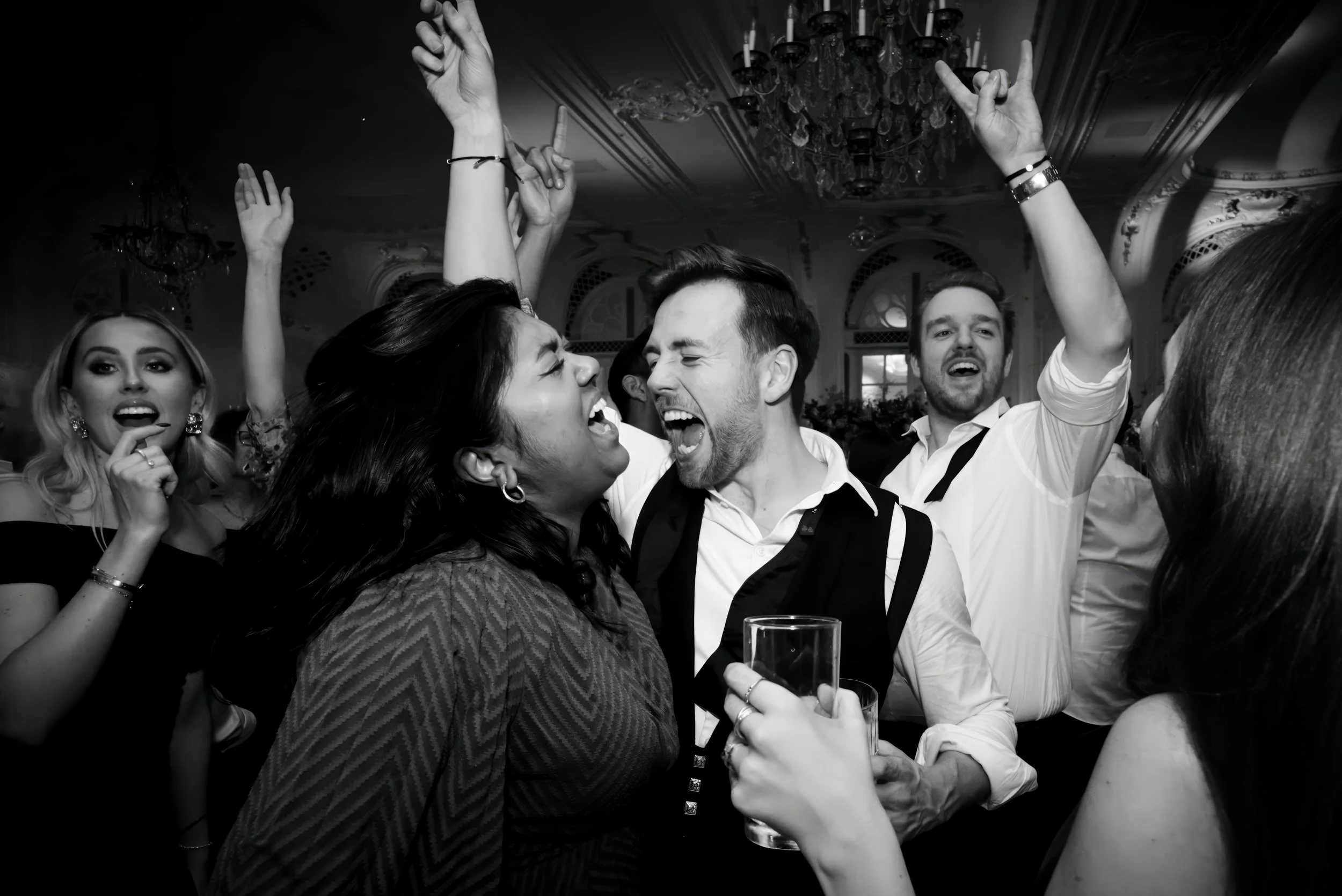 People dancing and singing at a party or celebration in an elegant room with chandeliers.