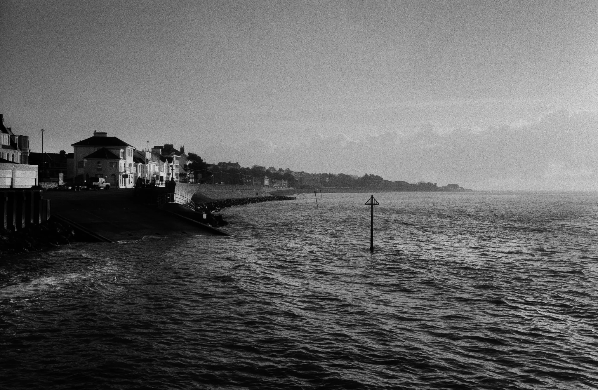A black and white photograph of a coastal scene with houses along the shoreline on the left side, and a calm water body extending to the horizon with a navigational aid post in the water.
