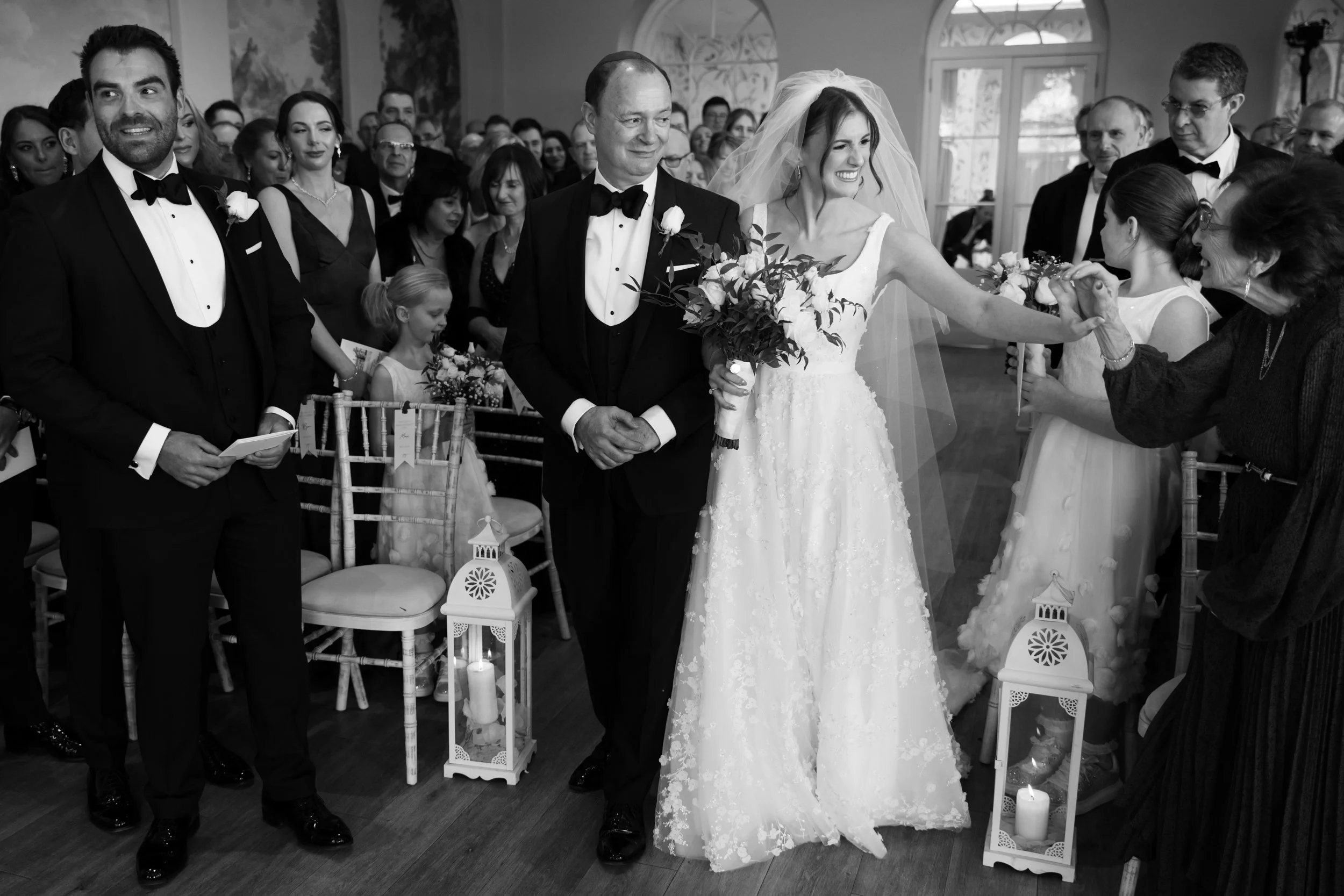 A bride in a white gown and veil holding a bouquet smiles as she is handed a flower by a woman during a wedding ceremony. The groom and other guests dressed in tuxedos and formal attire stand around in a decorated indoor venue with chairs, lanterns, 