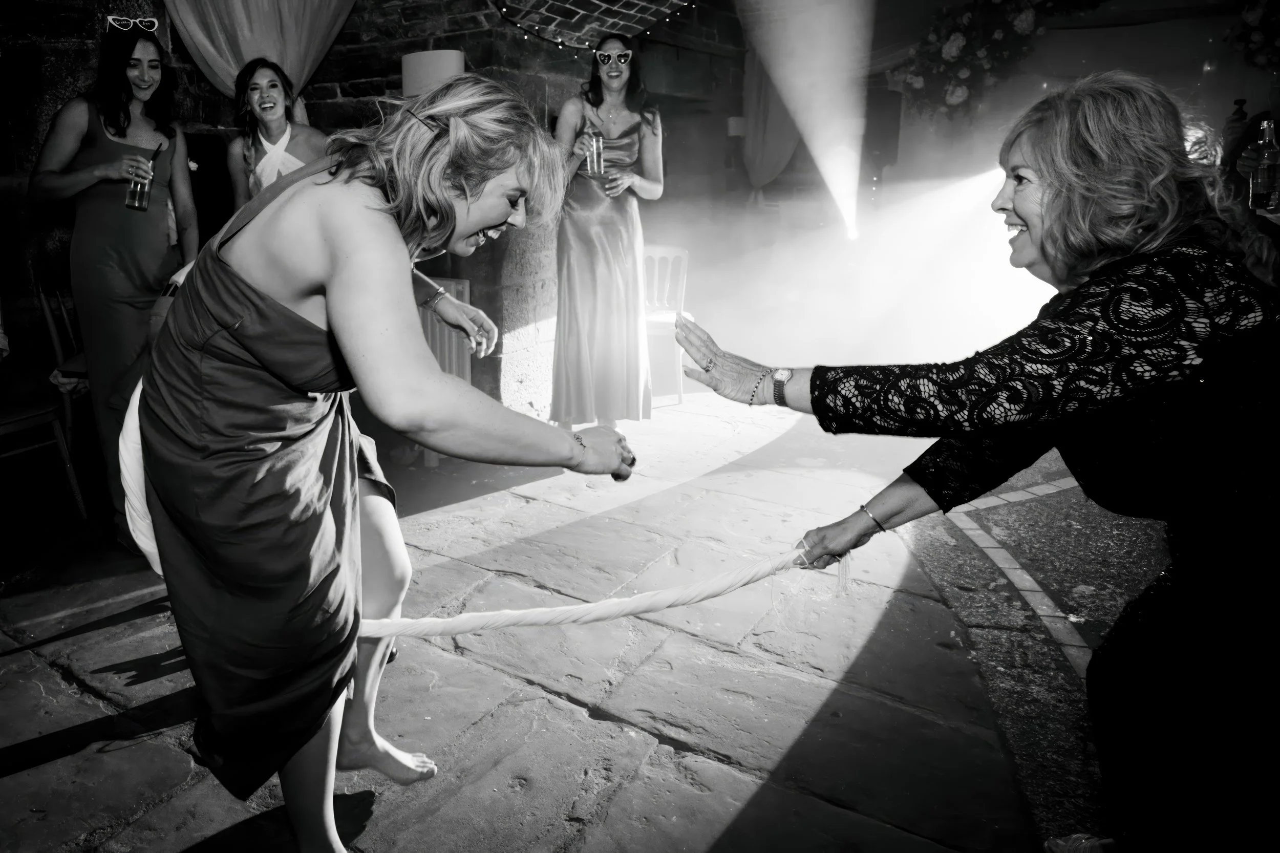 Women playing tug of war at a party or celebration, with others watching and smiling, black and white photo.