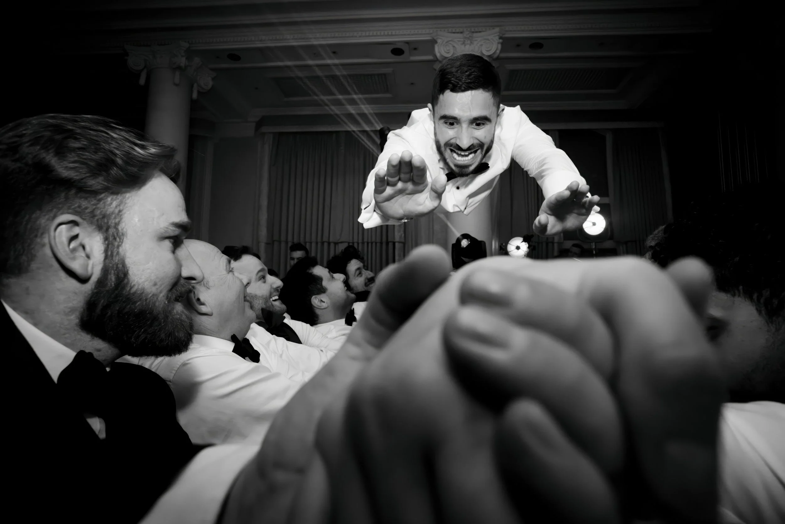 A man in a white shirt and bow tie appears to be flying or jumping above a row of people dressed formally, with the scene captured in black and white at what looks like a celebration or formal event.