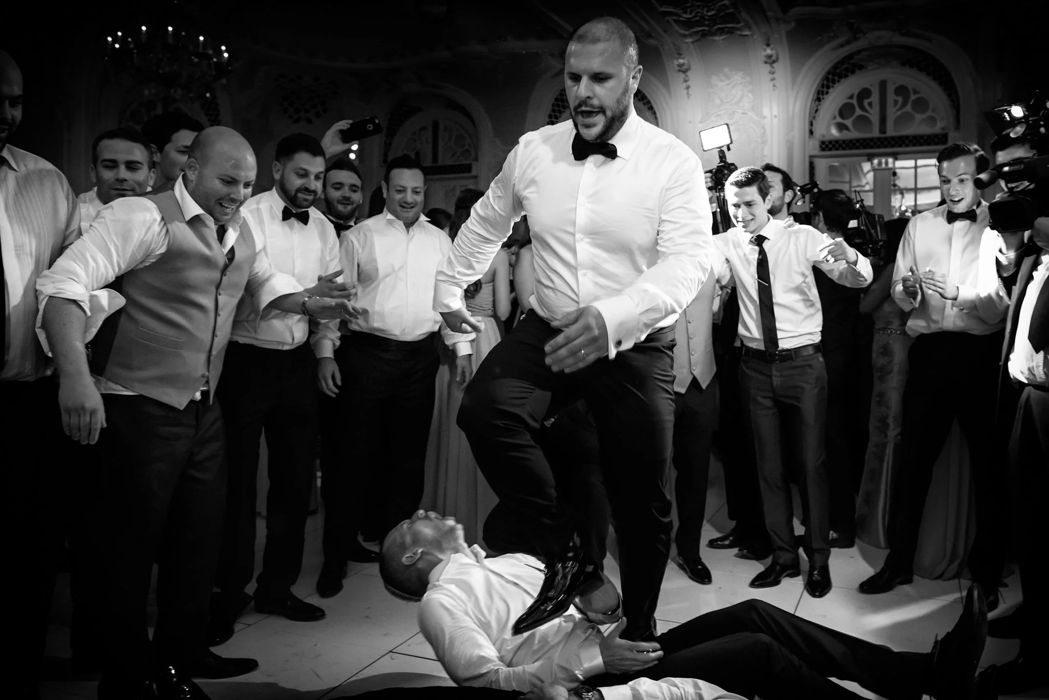 A group of men in formal attire dancing at an event, with one man standing over another lying on the floor, surrounded by onlookers.