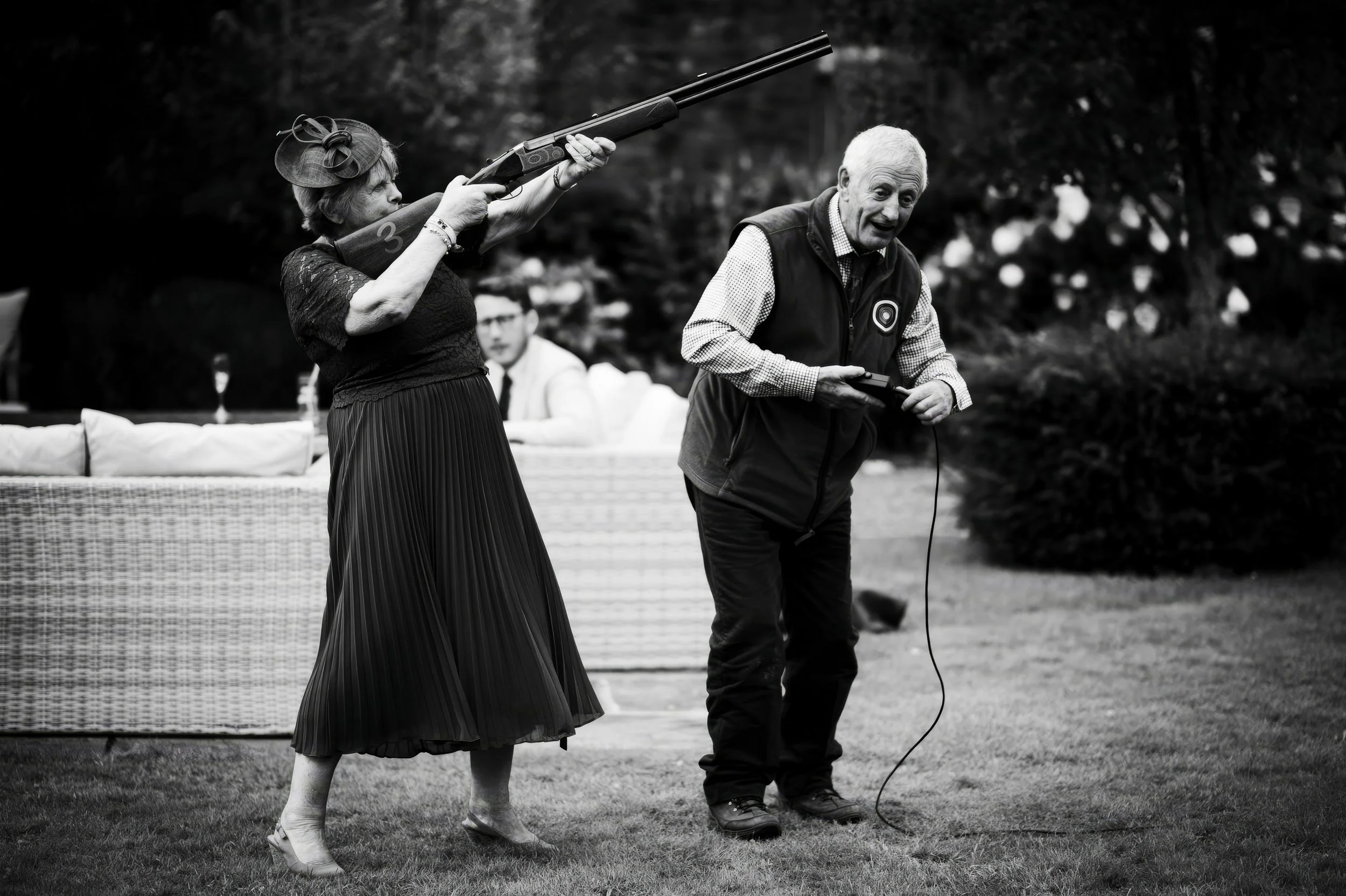 An older woman aiming a rifle, possibly in a shooting or hunting activity, while a man holding a device, perhaps a remote or a microphone, stands nearby on a grassy outdoor area. There is a person in the background sitting on a wicker couch.