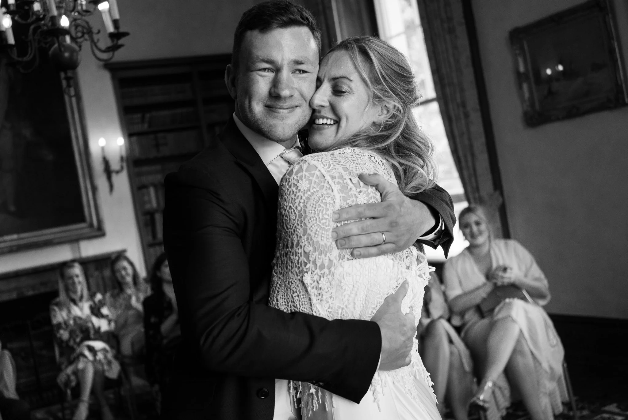 A black and white photo of a couple hugging, dressed in wedding attire, with smiling women seated in the background