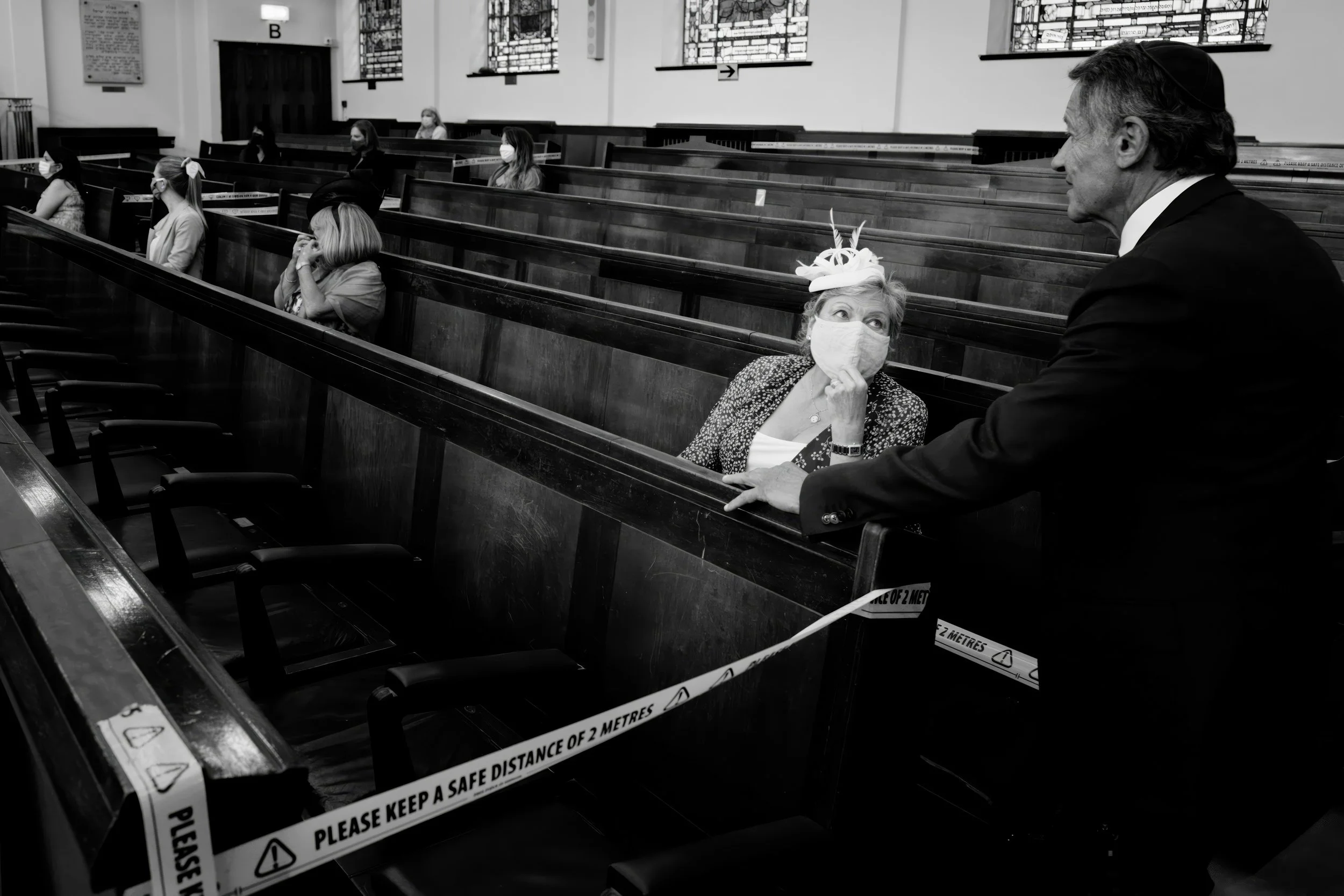 A woman in a hat and face mask sitting in a church pew, listening to a man in a suit speaking. Social distancing markers on the pews indicate COVID-19 precautions.