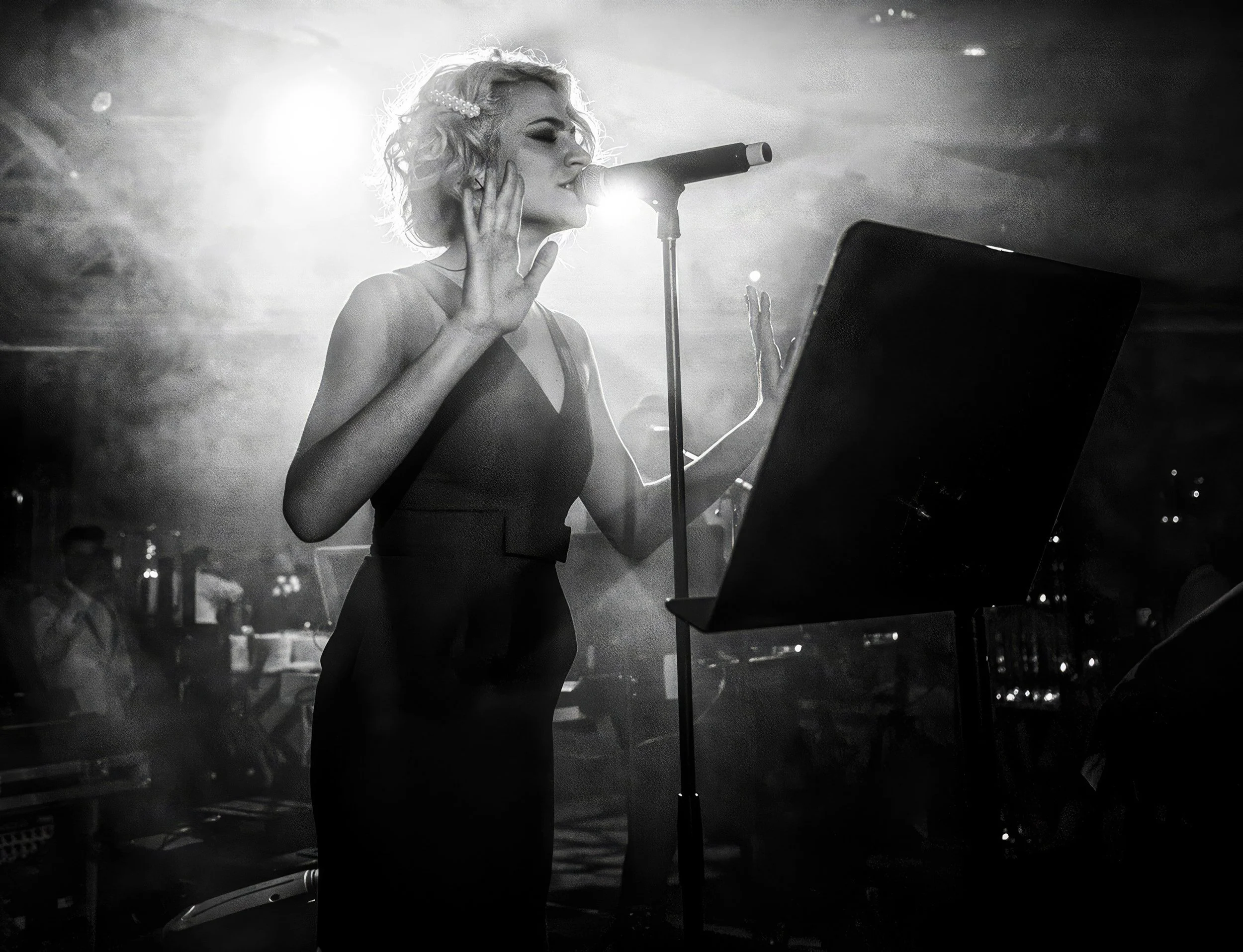 A woman with short, wavy blonde hair performing on stage, singing into a microphone, with stage lighting creating a dramatic effect.