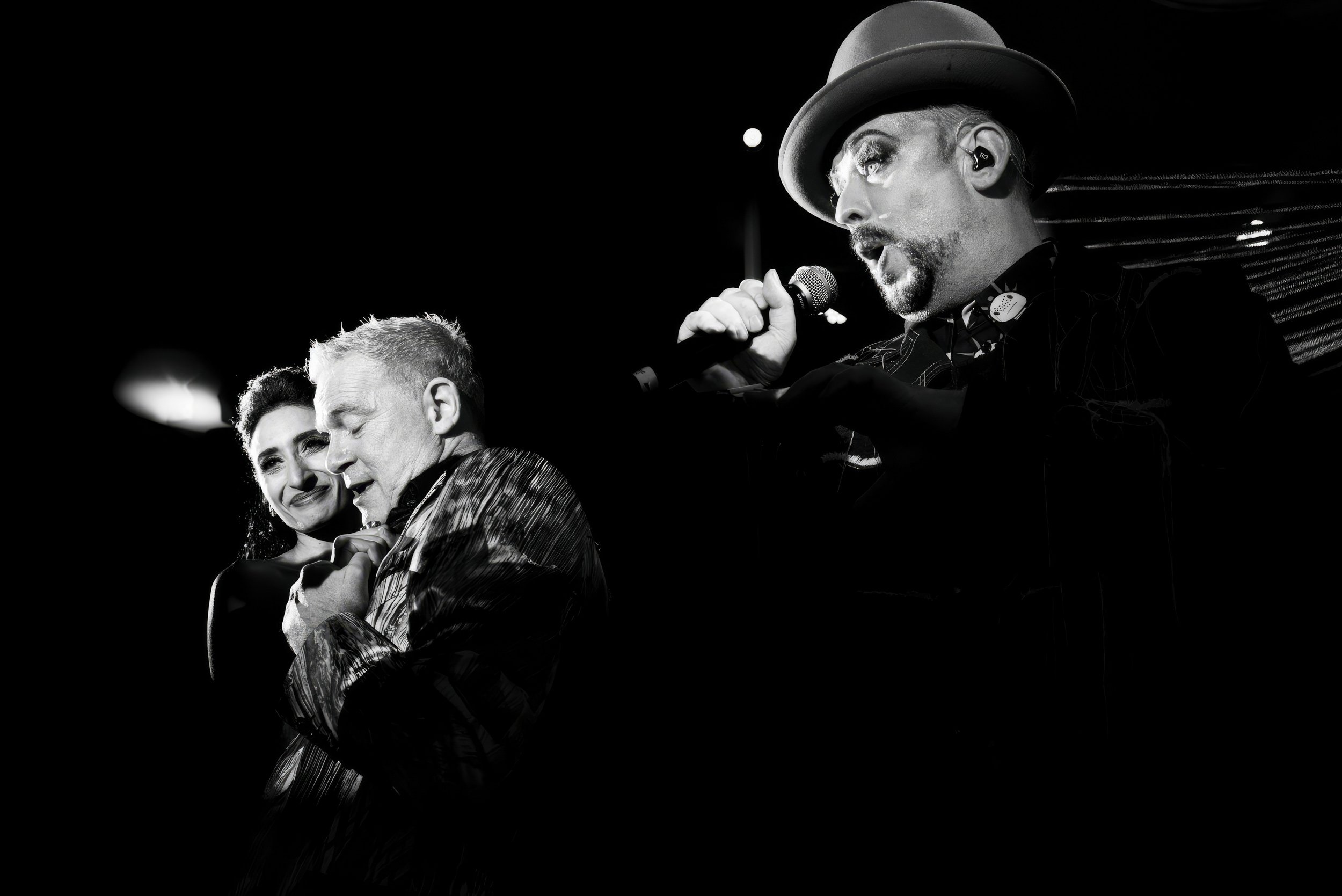 Black and white photo of a performance featuring a woman, an older man, and a singer. The woman and older man are close together, with the woman holding the man's hands, and the singer is singing into a microphone and wearing a hat.
