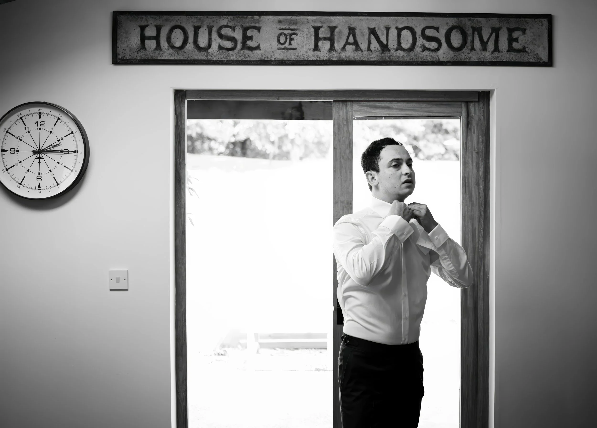 A man in a white dress shirt is adjusting his collar in front of a sliding glass door, with a clock on the wall and a sign reading 'House of Handsome' above him.