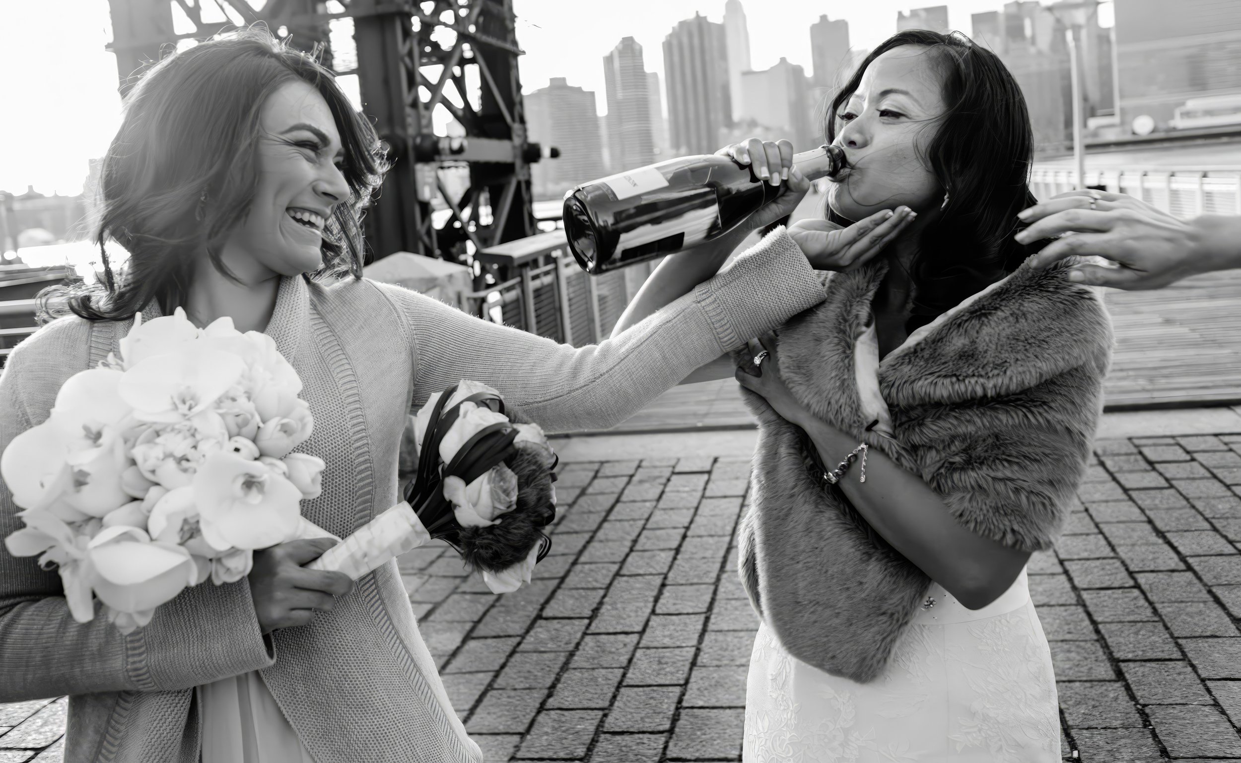 A woman in a wedding dress receiving a bottle of champagne from another woman holding a bouquet, with both women smiling outdoors in an urban setting.