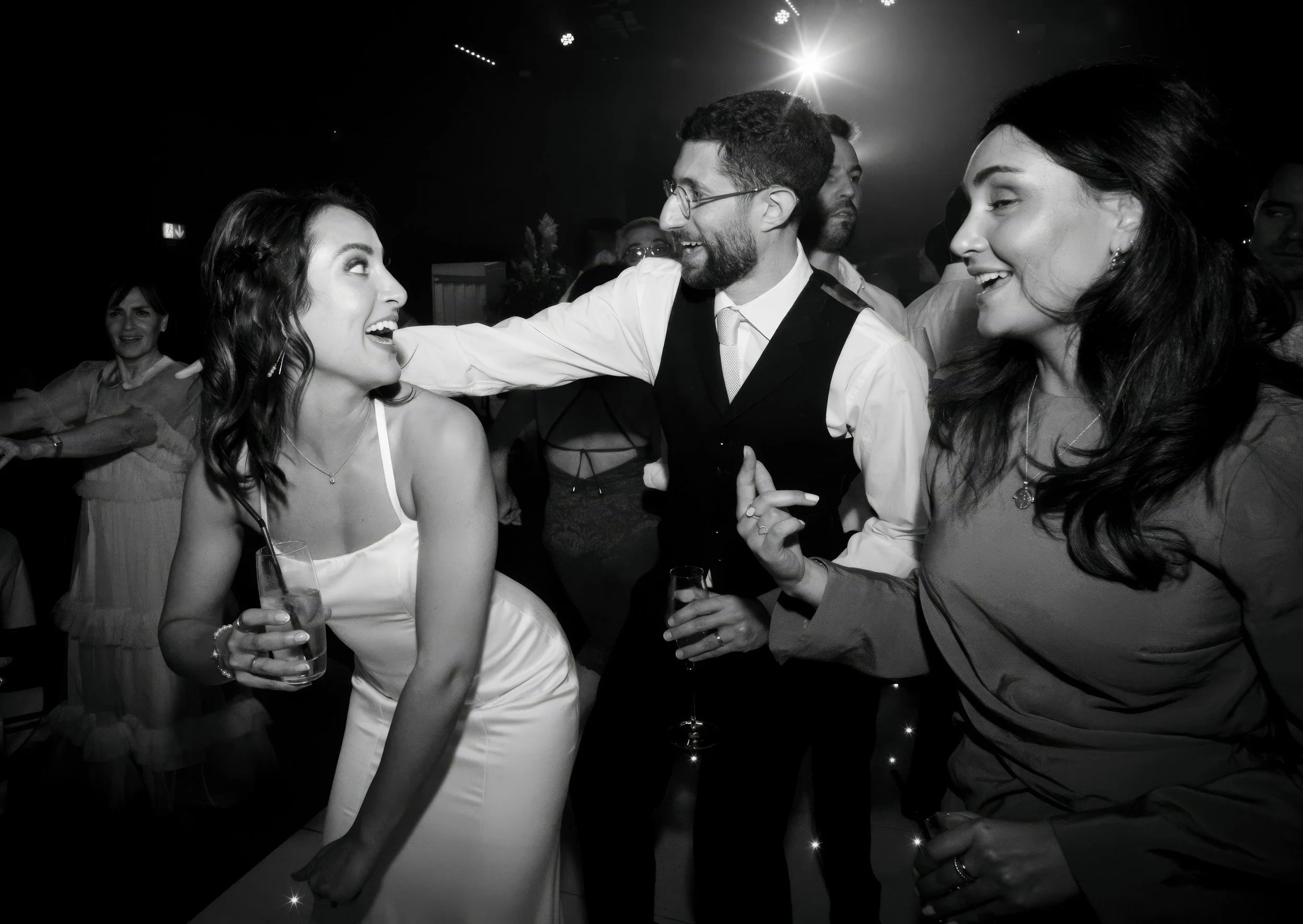 People dancing and socializing at a wedding reception, with a woman in a white dress holding a drink, smiling as she interacts with a man in a black vest, and a woman in a dark top smiling nearby, in a festive setting with a spotlight overhead.
