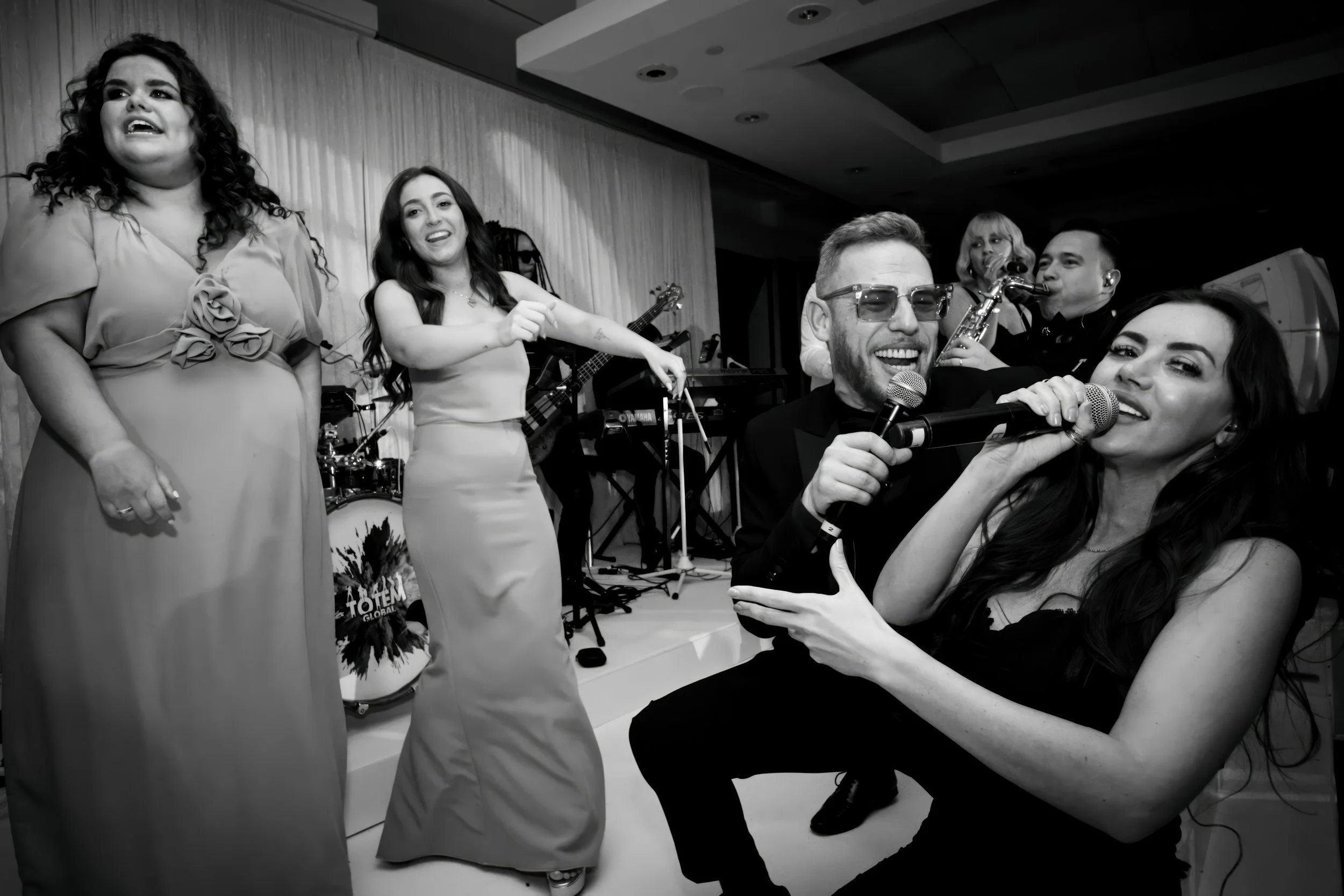 A group of people singing and performing on stage, including two women in dresses, a man with sunglasses singing into a microphone, a woman with long dark hair also singing, and a band playing instruments in the background.