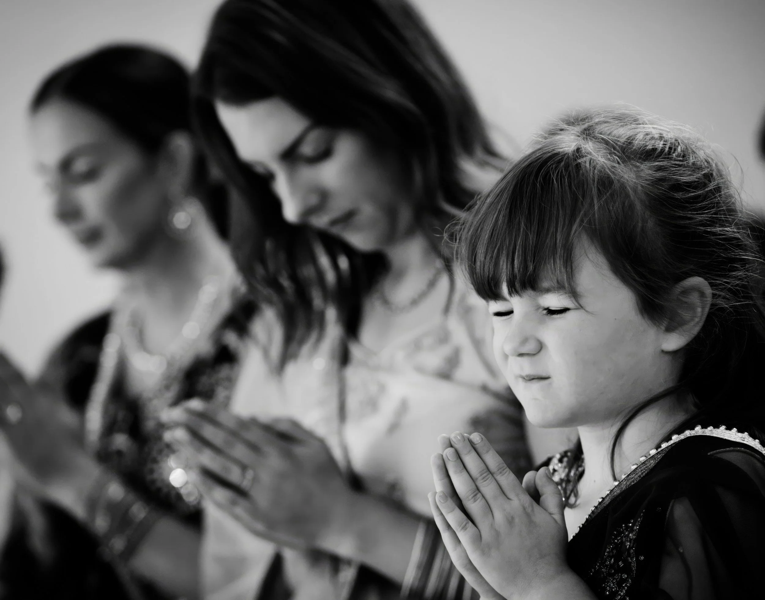 A young girl with dark hair and bangs, eyes closed, praying with hands pressed together, with two women in the background, also praying, seen in black and white.