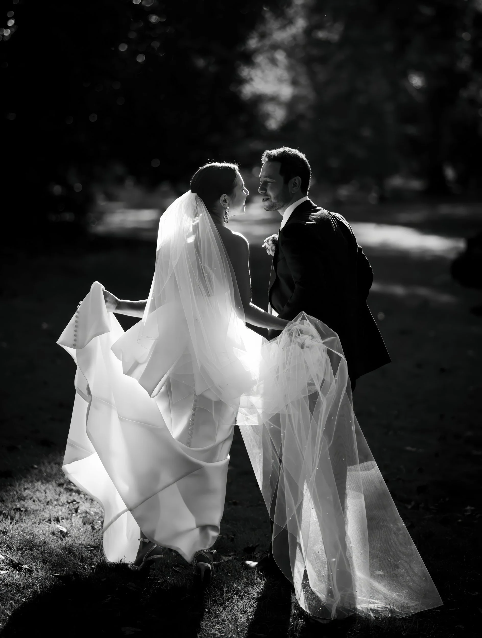 Black and white photo of a bride and groom holding hands, standing outdoors, facing each other, with trees in the background.