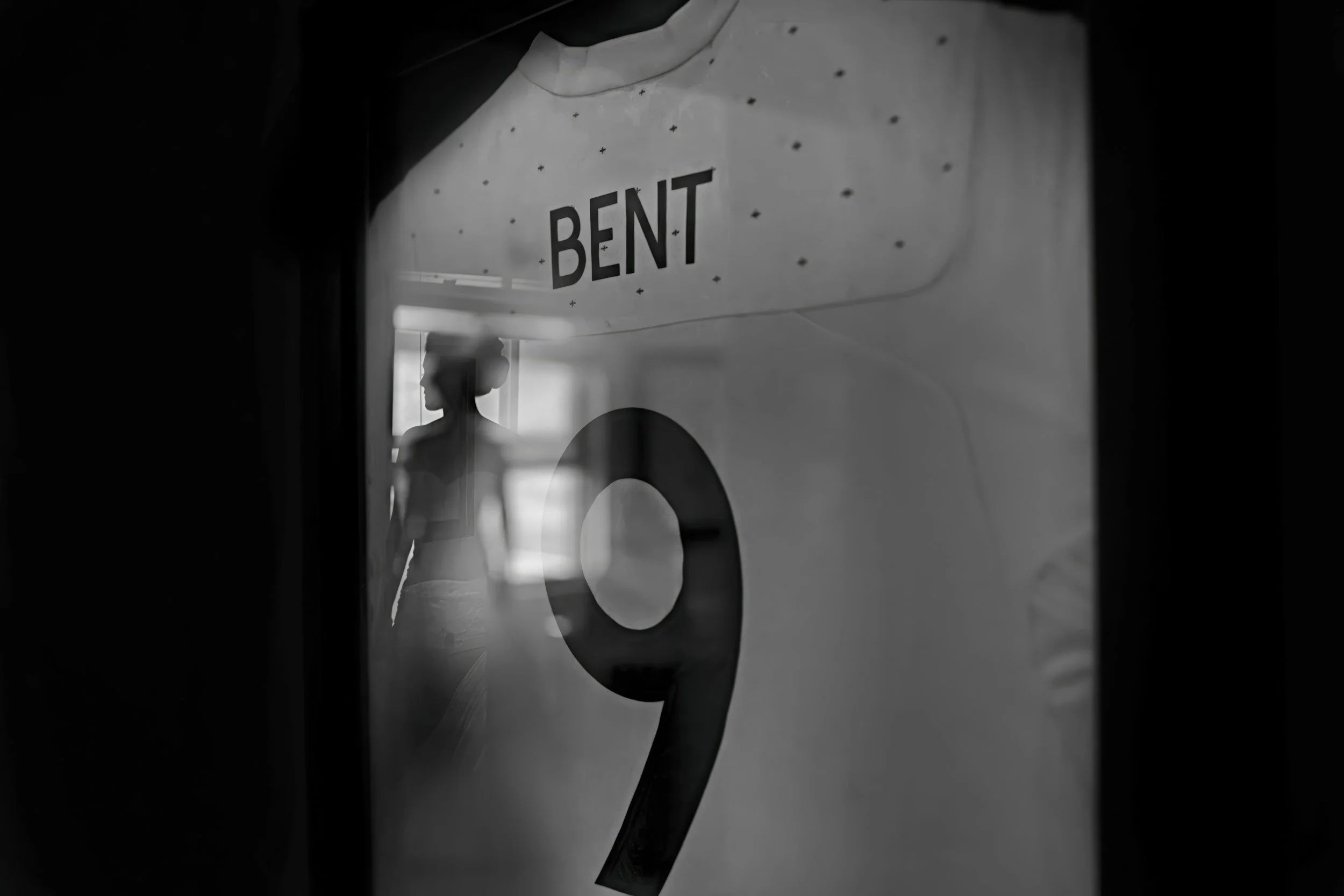 Silhouette of a woman with a bun hairstyle, standing inside a room, reflected on a glass surface. The photo is in black and white, with a large number 9 and the word 'BENT' visible in the foreground, likely on a poster or sign.