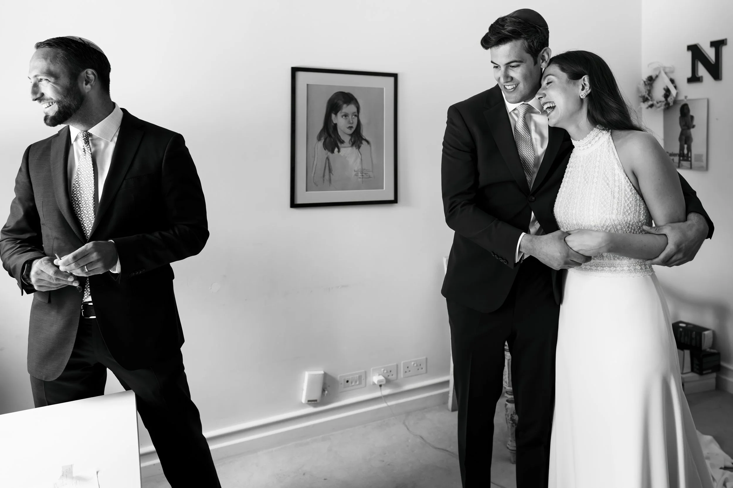 A black and white photo of a man and woman in wedding attire laughing and embracing, standing in a room with a framed portrait of a young girl on the wall.