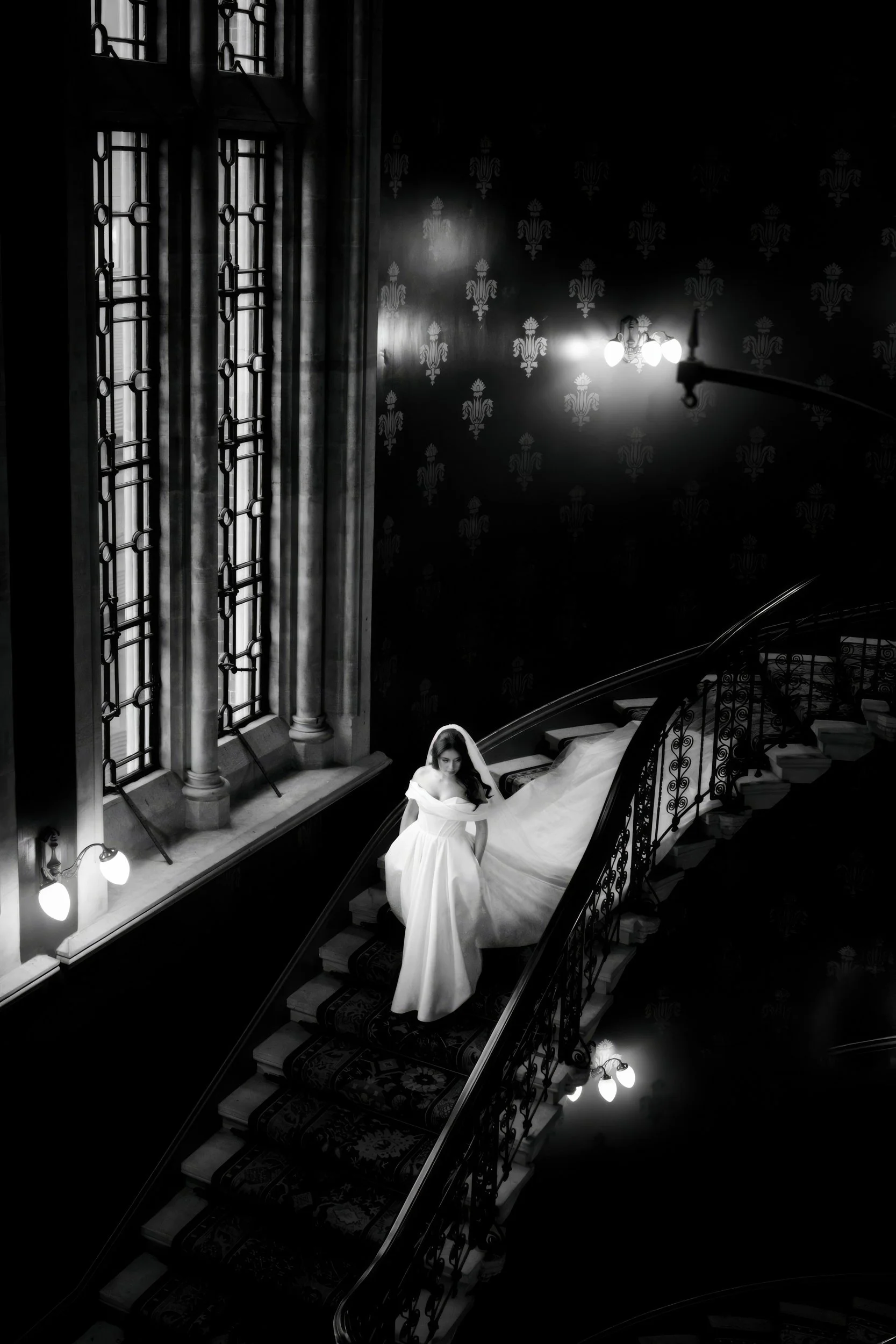 A woman in a wedding dress standing on a grand staircase inside a dimly lit, elegant building with large stained glass windows and ornate iron railings, illuminated by soft lighting.