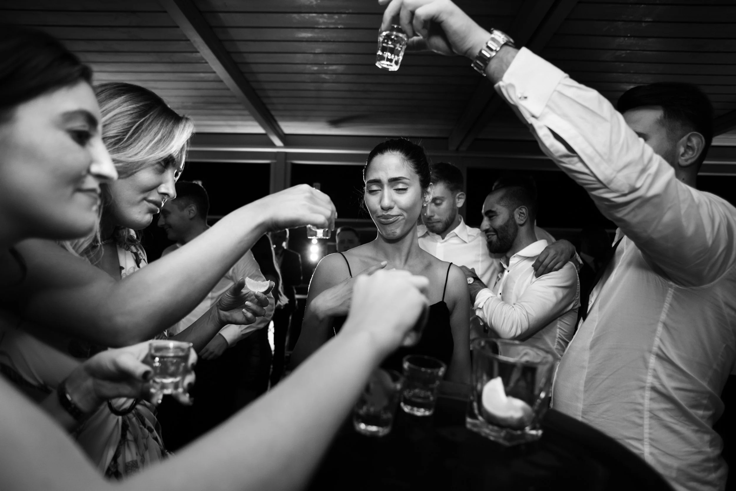 Group of people at a party holding drinks, with one woman in the center making a face.
