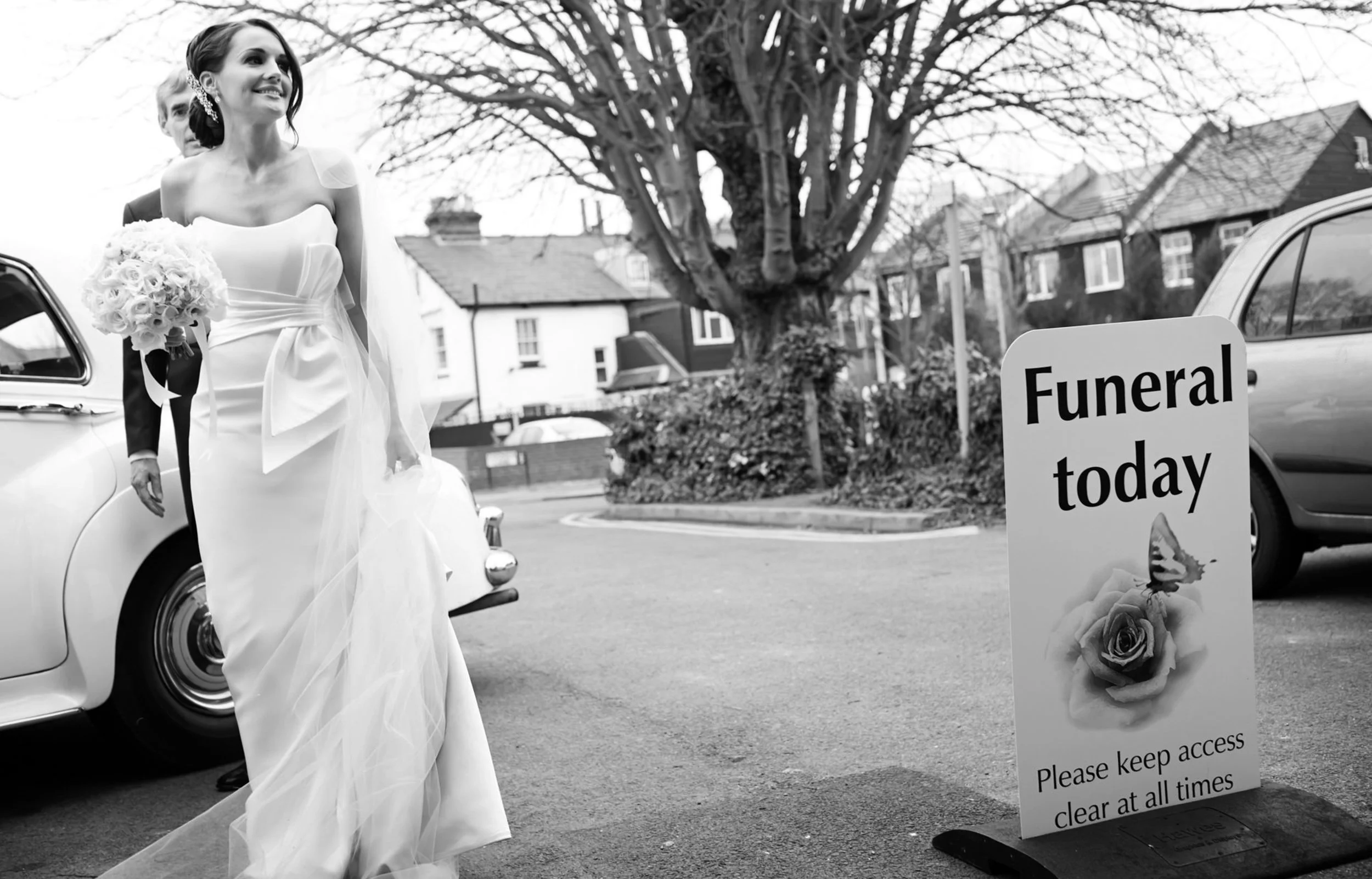 A bride in a wedding dress holding a bouquet, standing next to a car on a street with a funeral sign that reads 'Funeral today' and instructions to keep access clear.