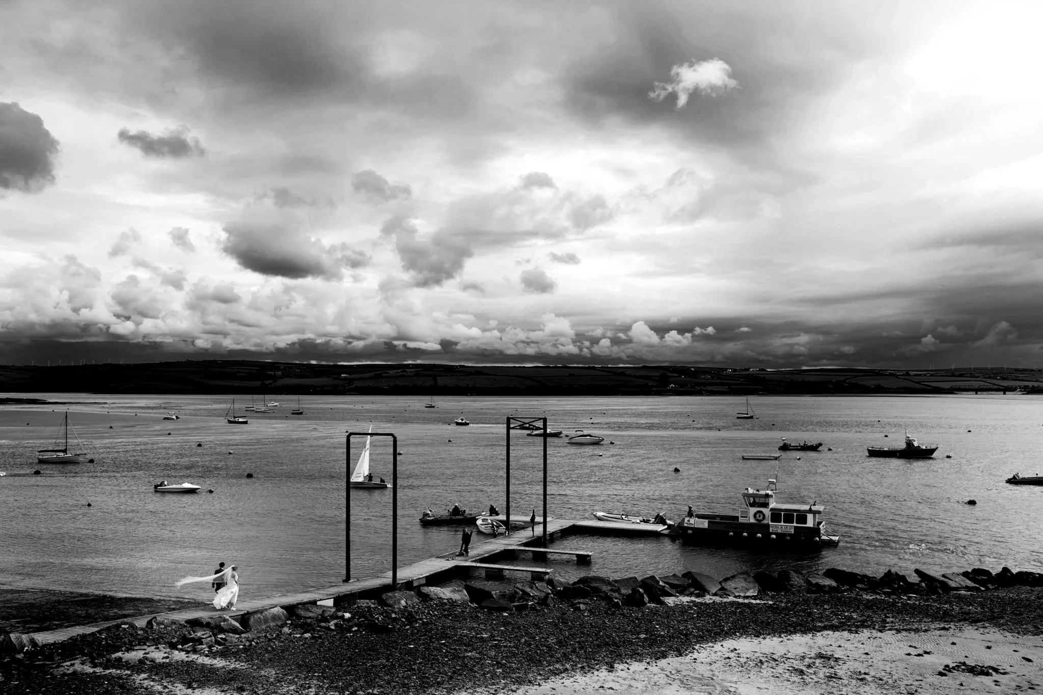 A black-and-white photo of a harbor with boats and a pier, two people in wedding attire walking on the dock, with dark, cloudy skies overhead.