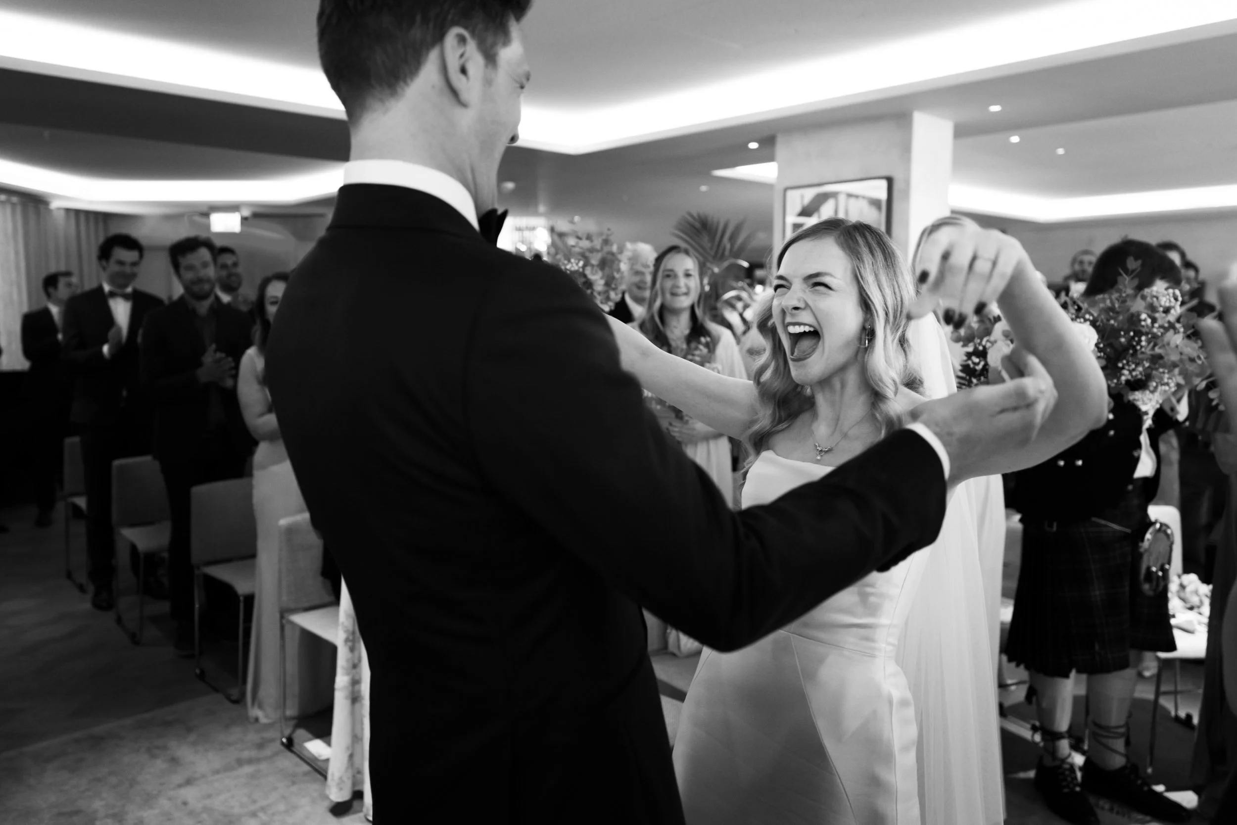A black and white photo of a bride and groom dancing at their wedding reception, surrounded by guests in a decorated banquet hall.