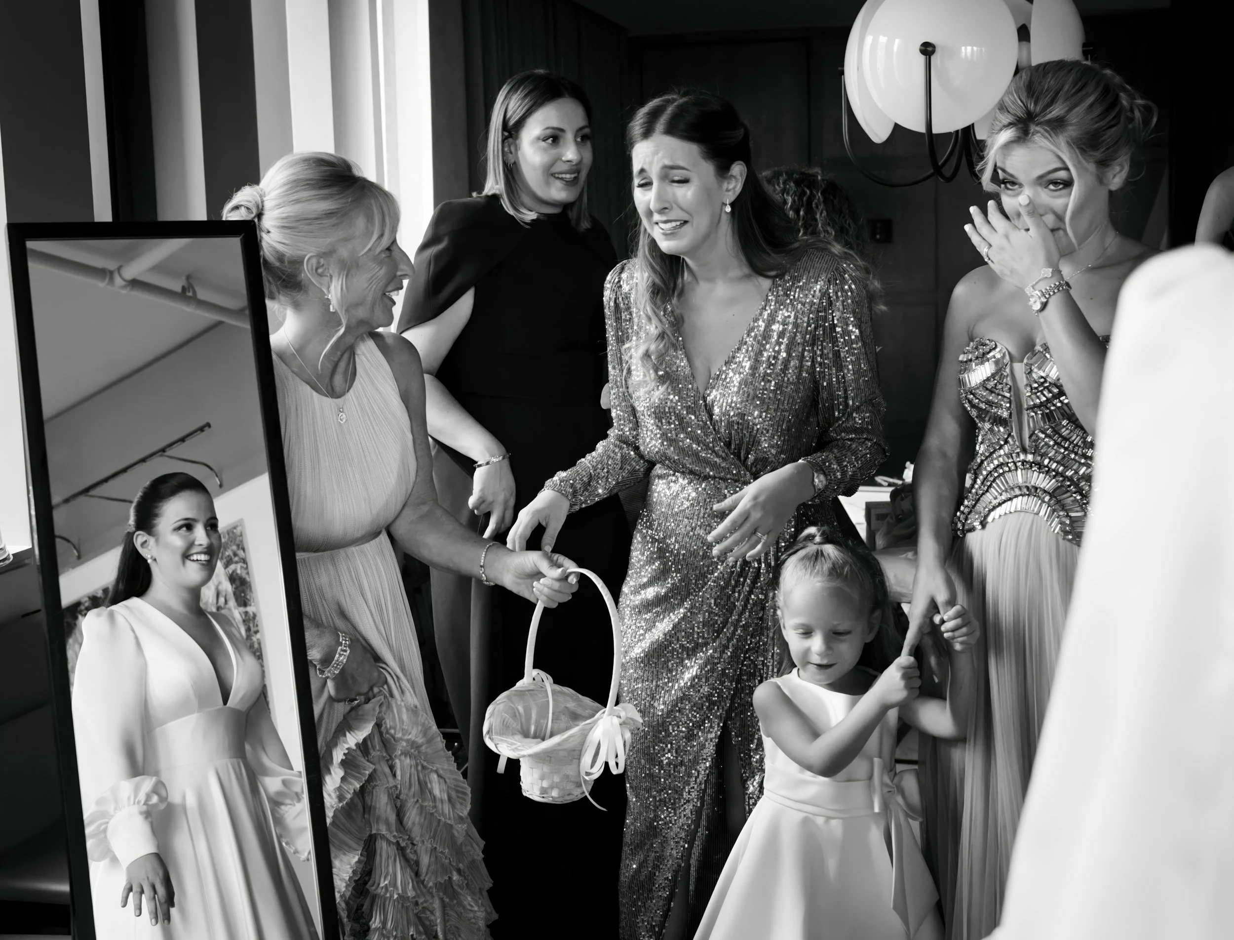 A group of women and a girl in elegant dresses reacting emotionally, with some smiling and one covering her mouth, with a mirror reflecting a woman in a white dress.
