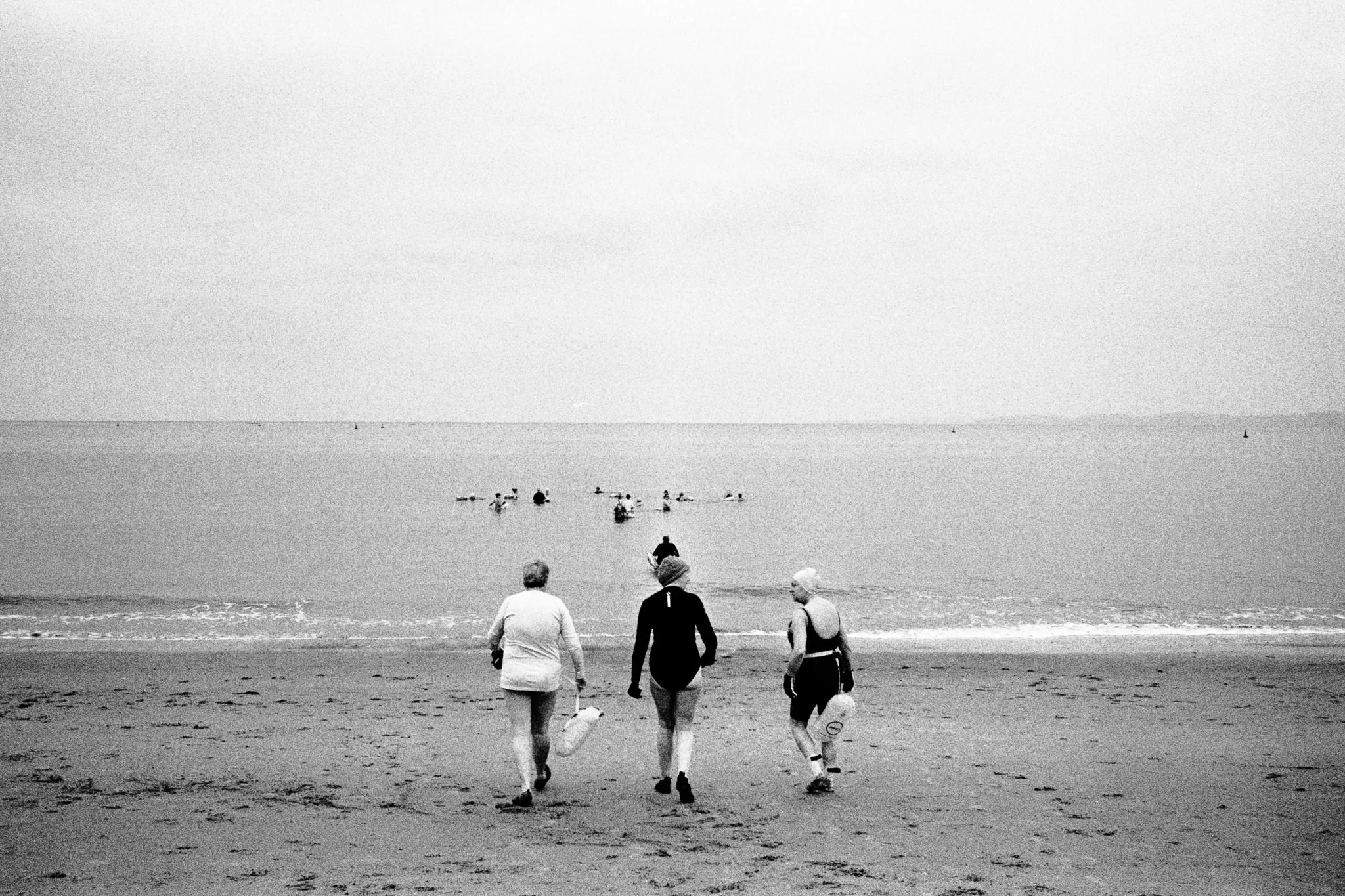 Three elderly women walking on a beach towards the water, with other people swimming and surfing in the ocean in the background.