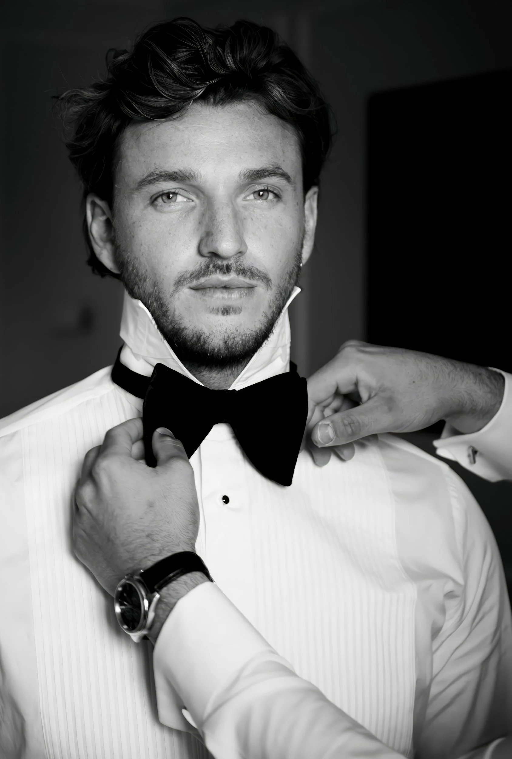 A young man with light skin and wavy dark hair is being helped to put on a black bow tie by another person. He is wearing a white dress shirt with a cufflink, and a watch on his left wrist. The setting is indoors, and the photo is in black and white.