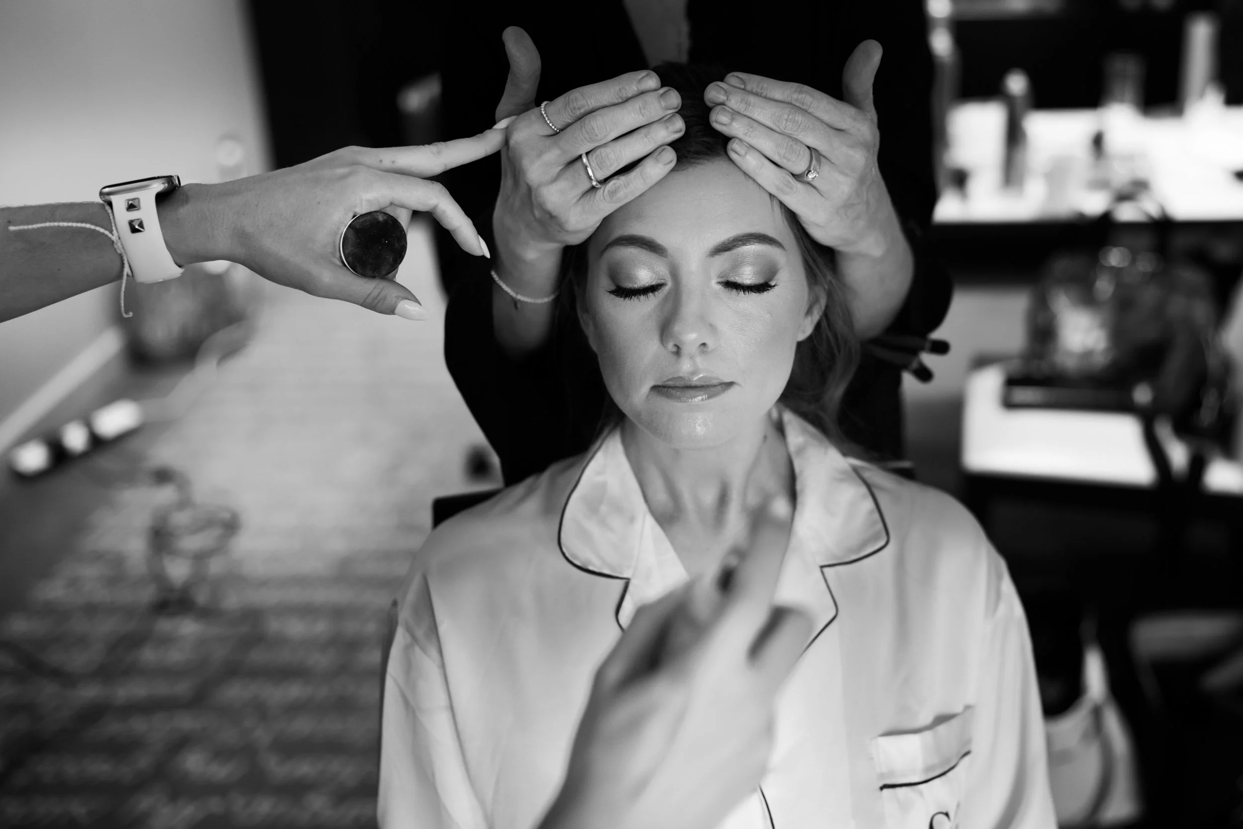 A woman with makeup and pajamas having her hair and makeup styled by a stylist behind her with hands on her head, while another person styles her face in a professional setting.
