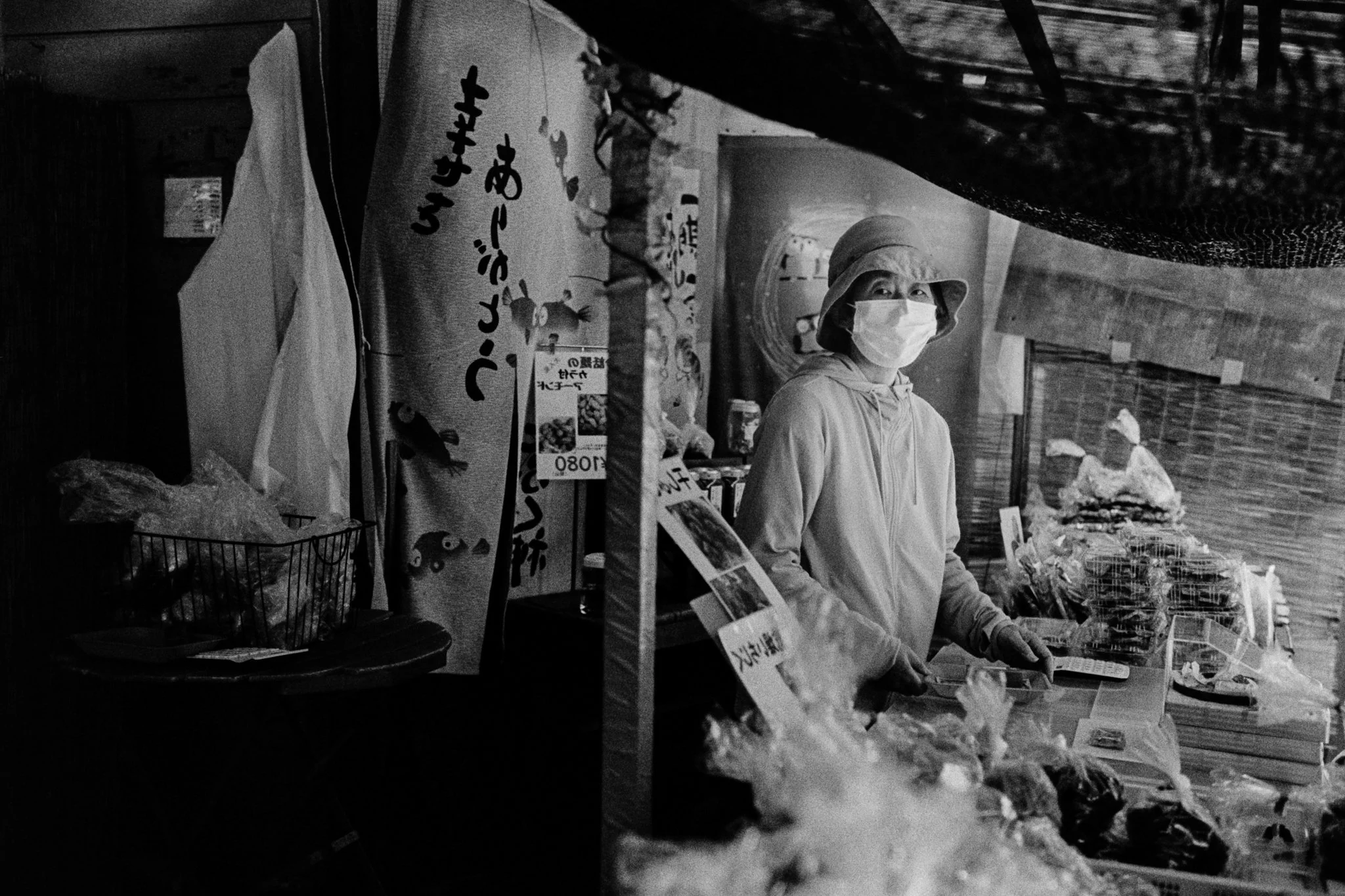 A person wearing a face mask and a bucket hat shopping at a market stand with various items, including packaged goods, on a table in front of them.