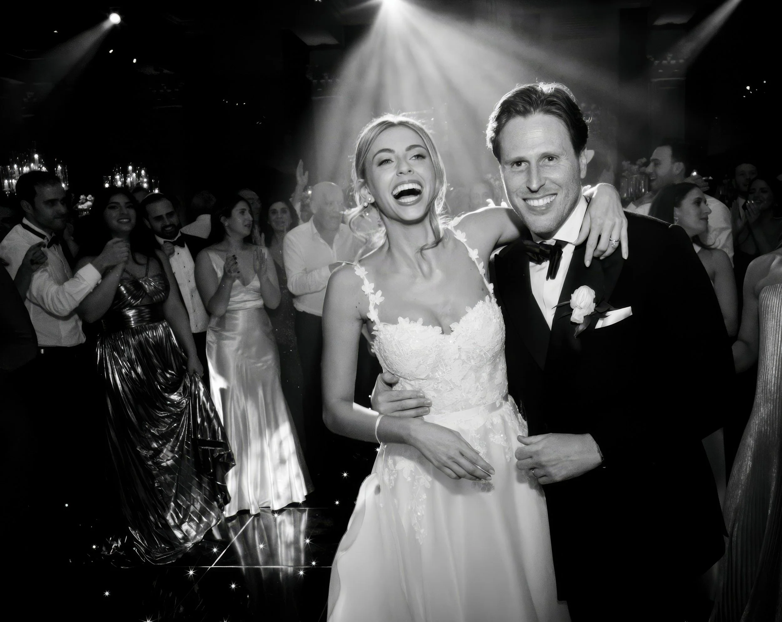 Black and white photo of a bride in a wedding dress and a groom in a tuxedo smiling and celebrating at a wedding reception with guests dancing and enjoying themselves in the background.