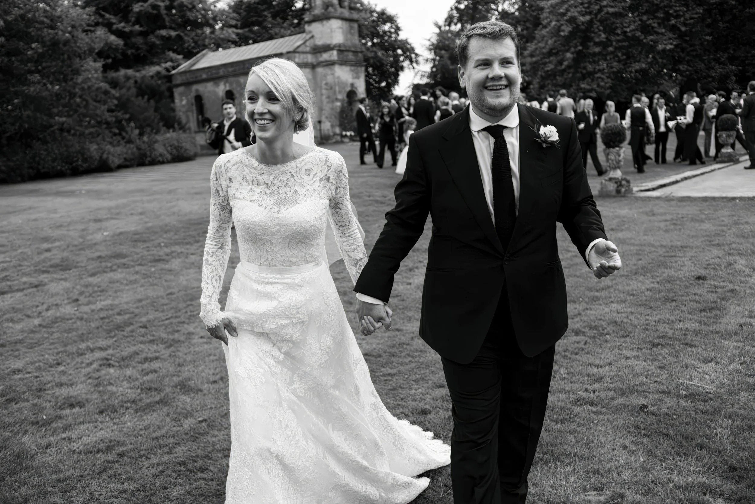 A black-and-white photo of a newlywed couple walking hand-in-hand outdoors, with a group of people in the background, possibly at a wedding reception or celebration.