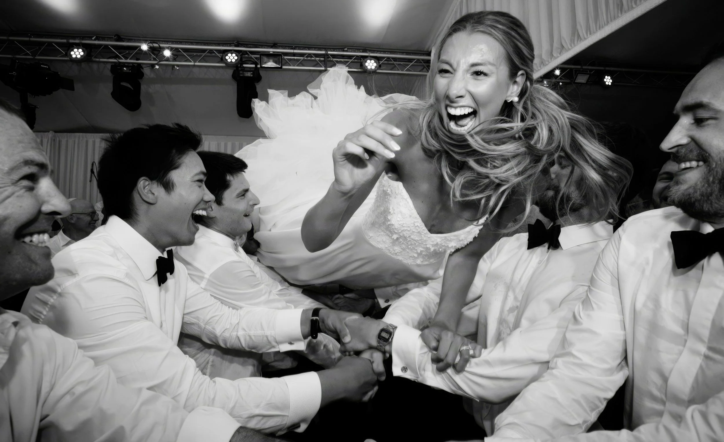A group of people in formal attire celebrating a wedding reception, with a woman in a wedding dress being lifted and laughing.